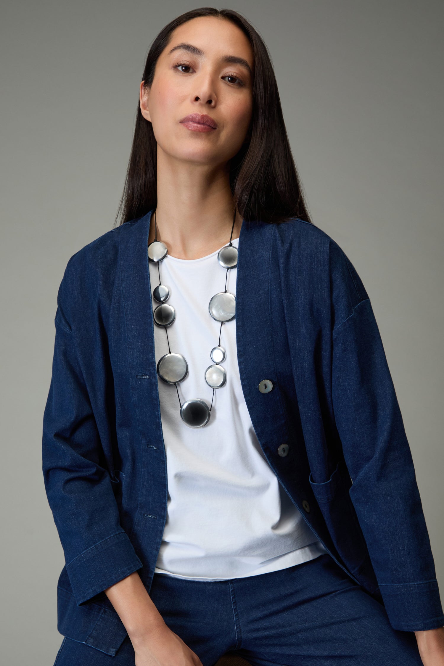 A woman with long dark hair wears a blue jacket over a white shirt and the Orbit Necklace, posing against a plain grey background.