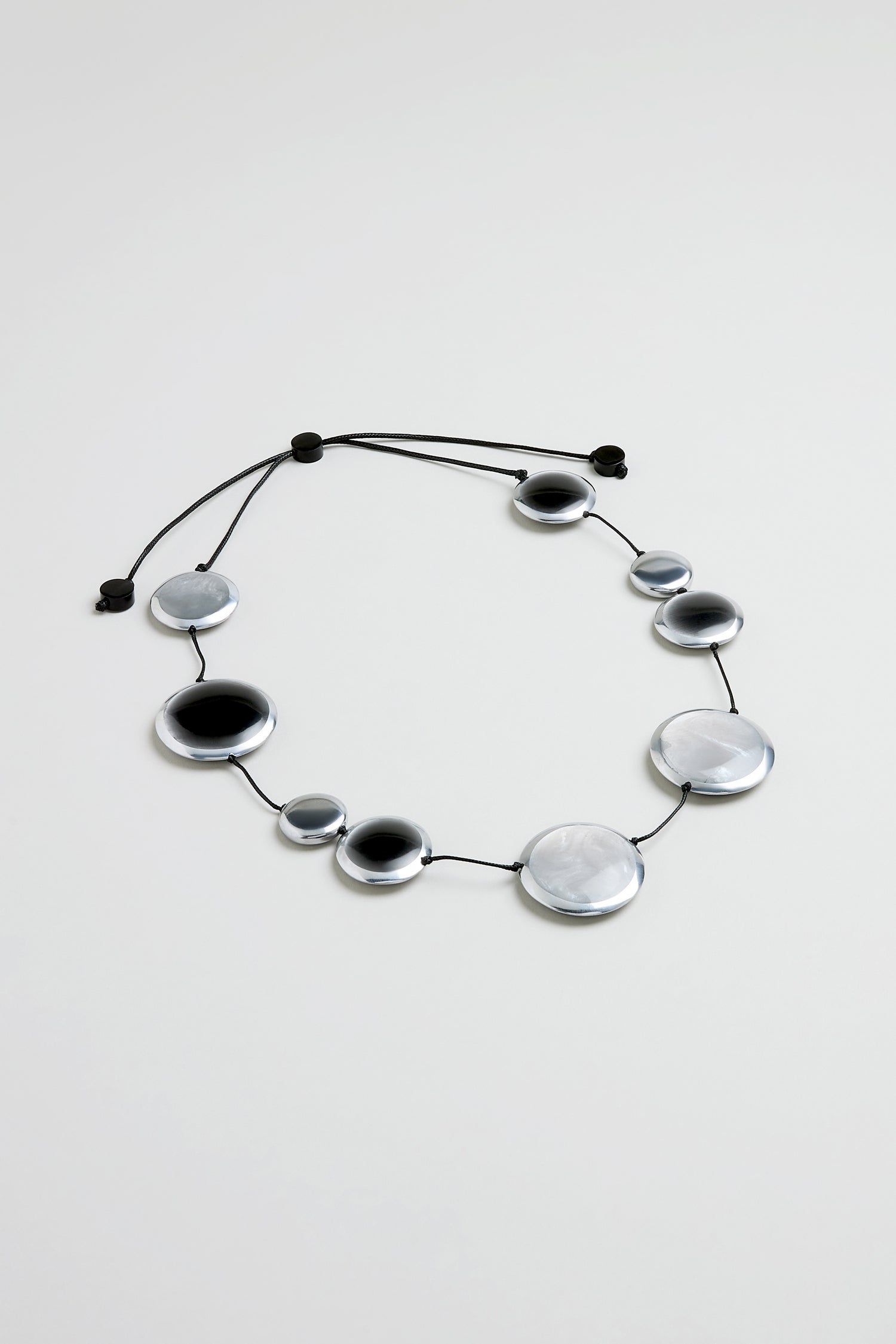 The Orbit Necklace features round silver and black disk-shaped beads of varying sizes on a black cord, highlighting modern craftsmanship.