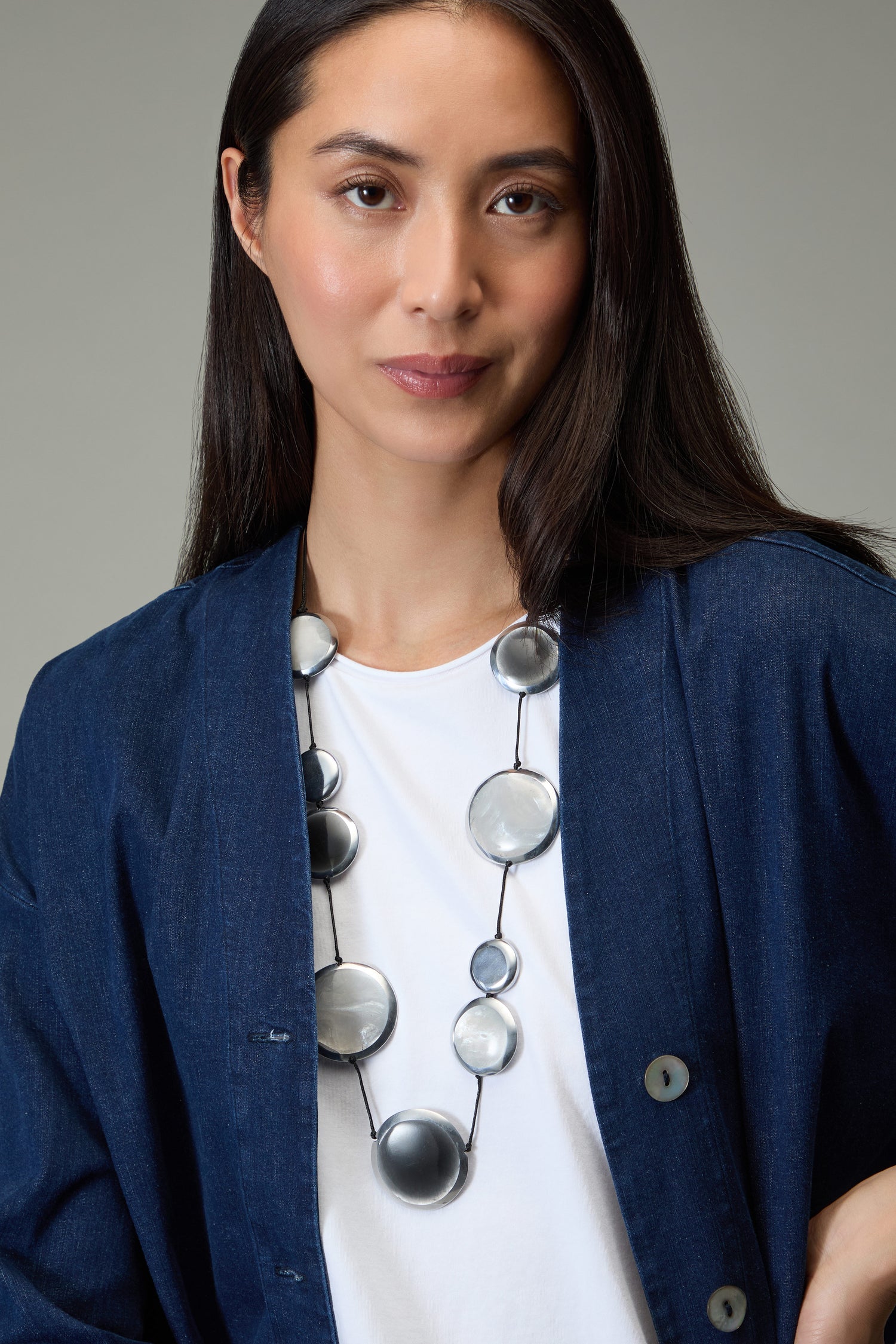A woman with long dark hair wears a blue jacket, white shirt, and the Orbit Necklace—posing against a plain background.