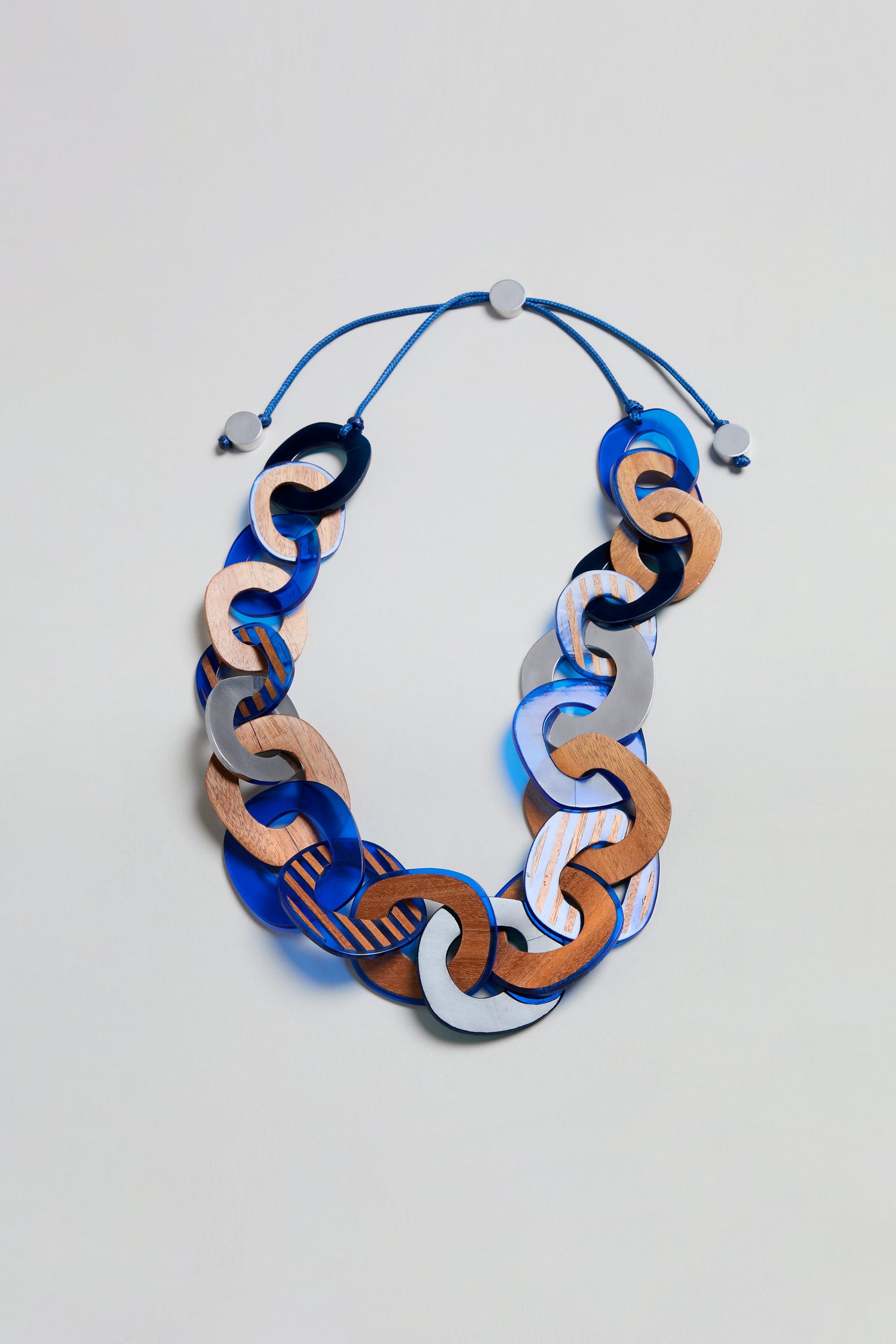 The Blue Stripe Wooden Resin Links Necklace features large interlocking links crafted from wood, blue resin, and silver-toned materials—some showcasing a distinctive striped wooden design. It is paired with a blue adjustable cord and circular fasteners, all displayed against a plain background.