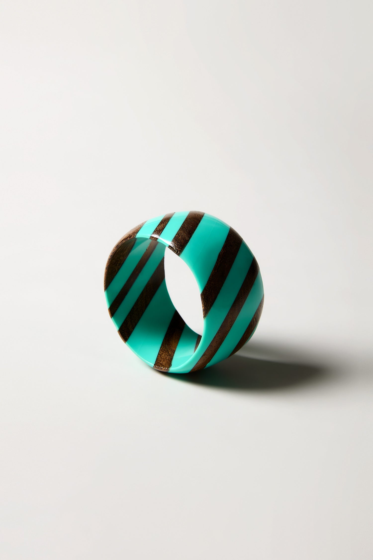 The Turquoise Wooden Stripe Bangle, handmade in Barcelona, showcases alternating turquoise and brown diagonal stripes and is displayed upright against a plain light background.