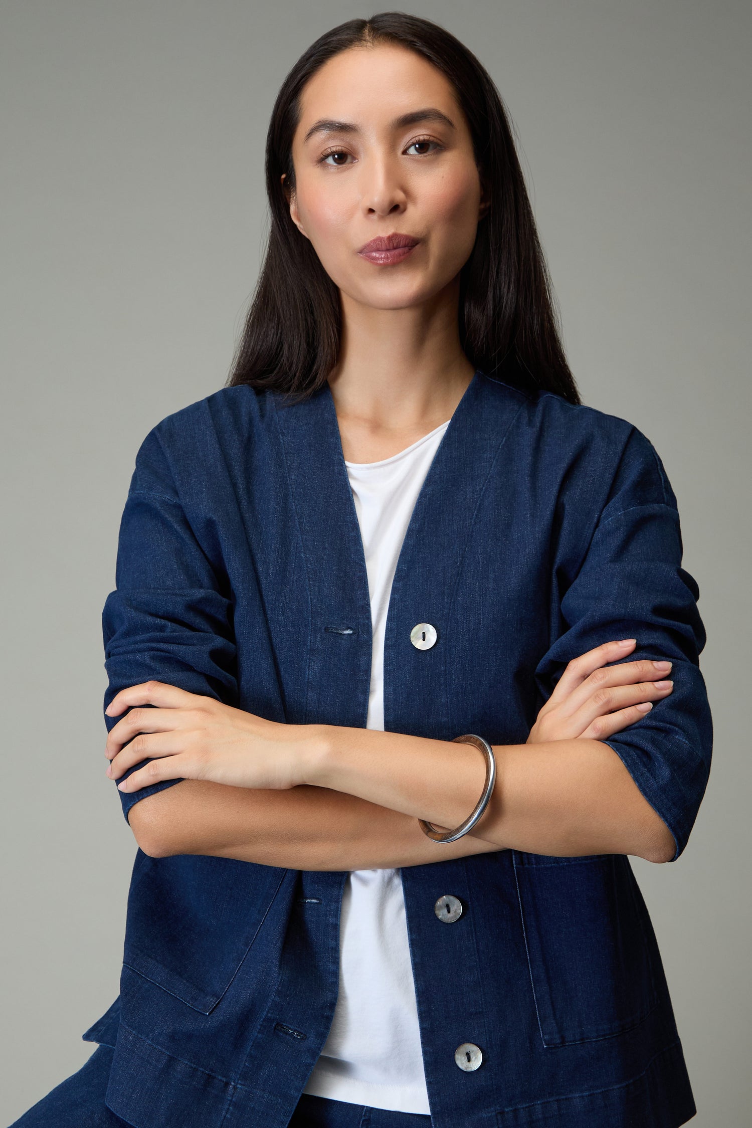 A woman with long dark hair, dressed in a blue jacket over a white shirt, stands with her arms crossed, wearing the Wooden Silver Fusion Bangle against a plain background.