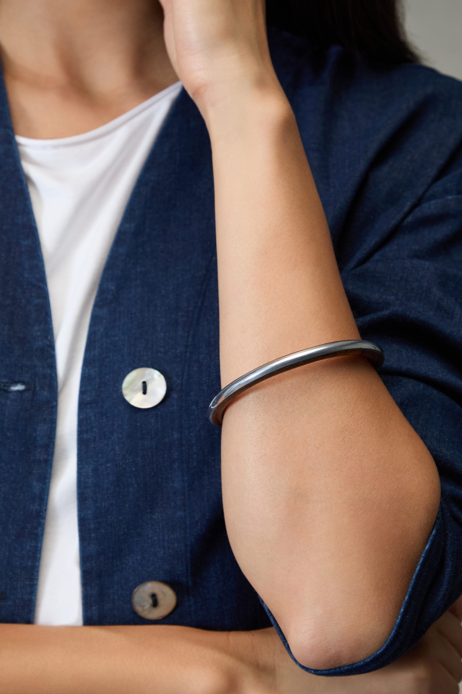 A person in a blue jacket and white shirt rests their hand on their face, displaying the Wooden Silver Fusion Bangle on their forearm.