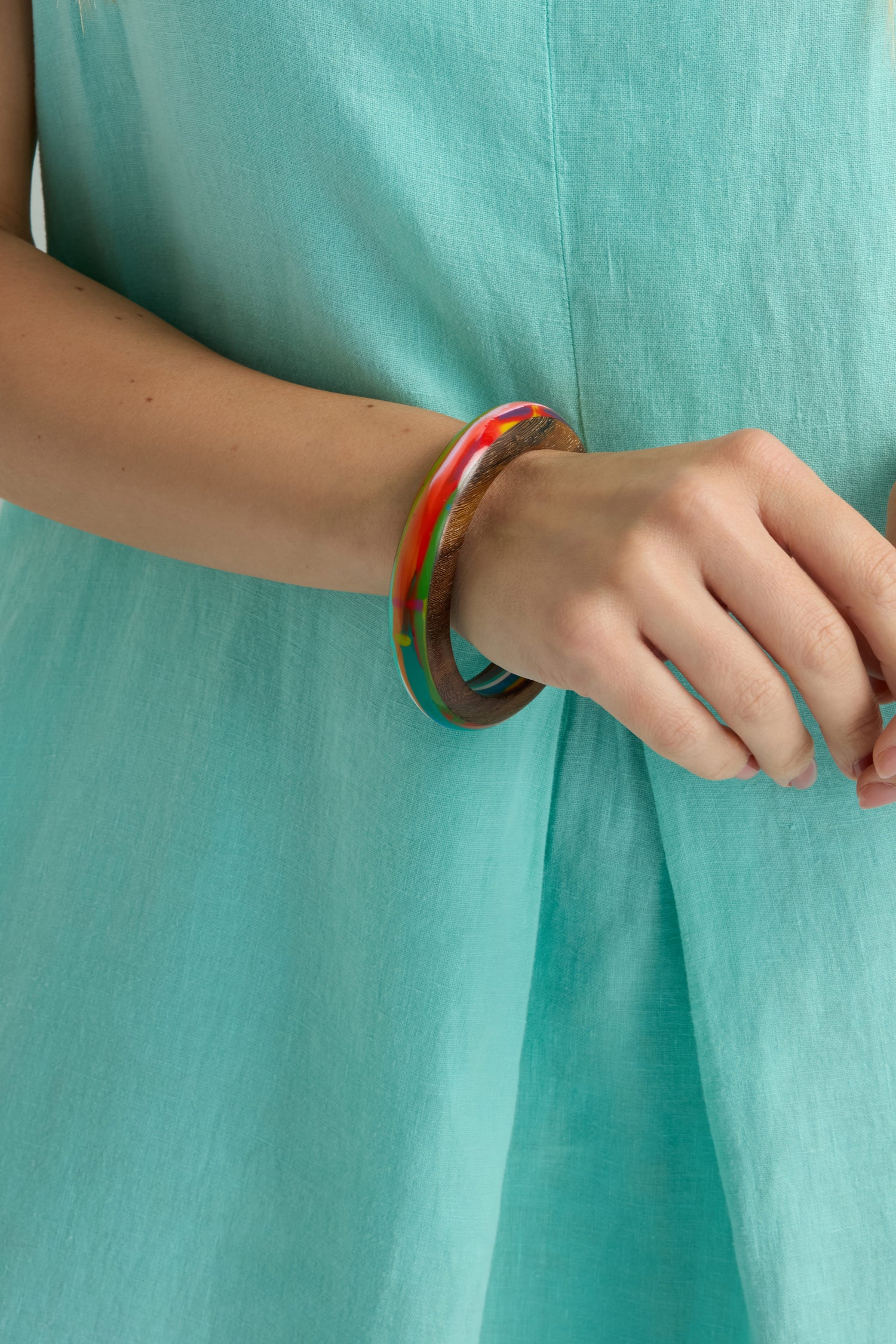 A person wearing a turquoise dress and two bracelets—one a polished wood bangle, the other the Flores Rainbow Bangle in vibrant multicoloured resin—on their wrist.