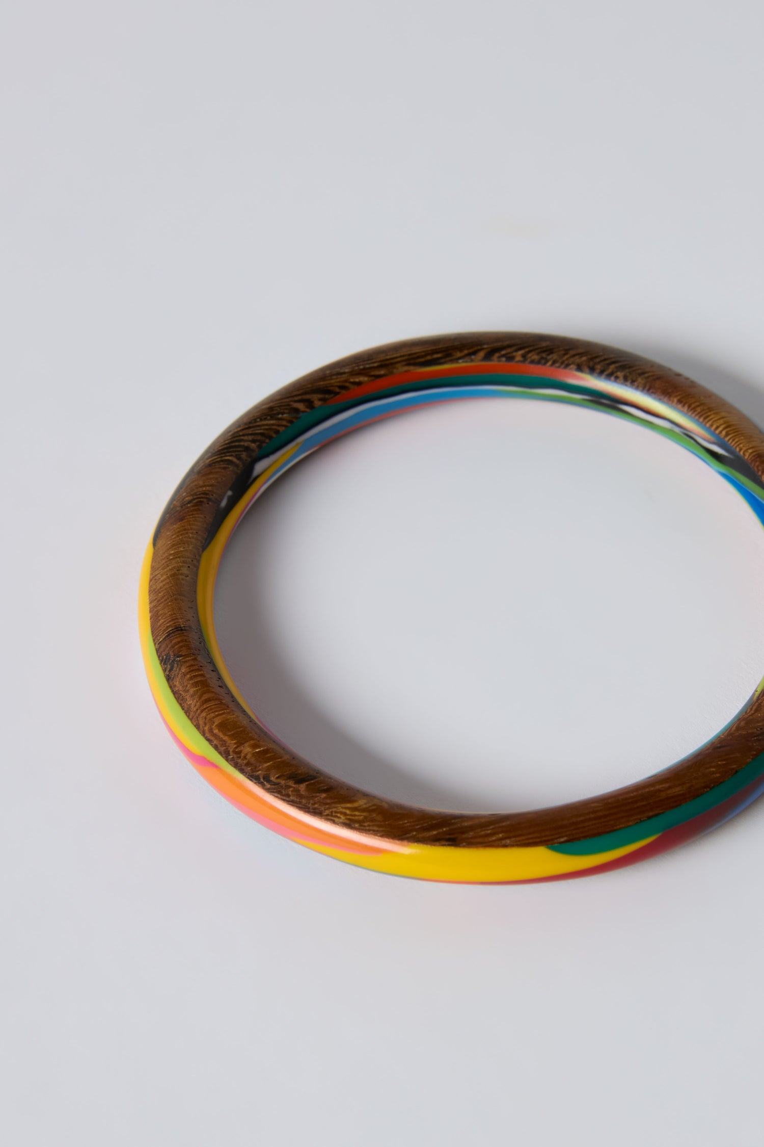 The Flores Rainbow Bangle is a round, sculptural bracelet with a polished wood-textured exterior and multicoloured resin sections in yellow, pink, green, and blue on a plain white background.