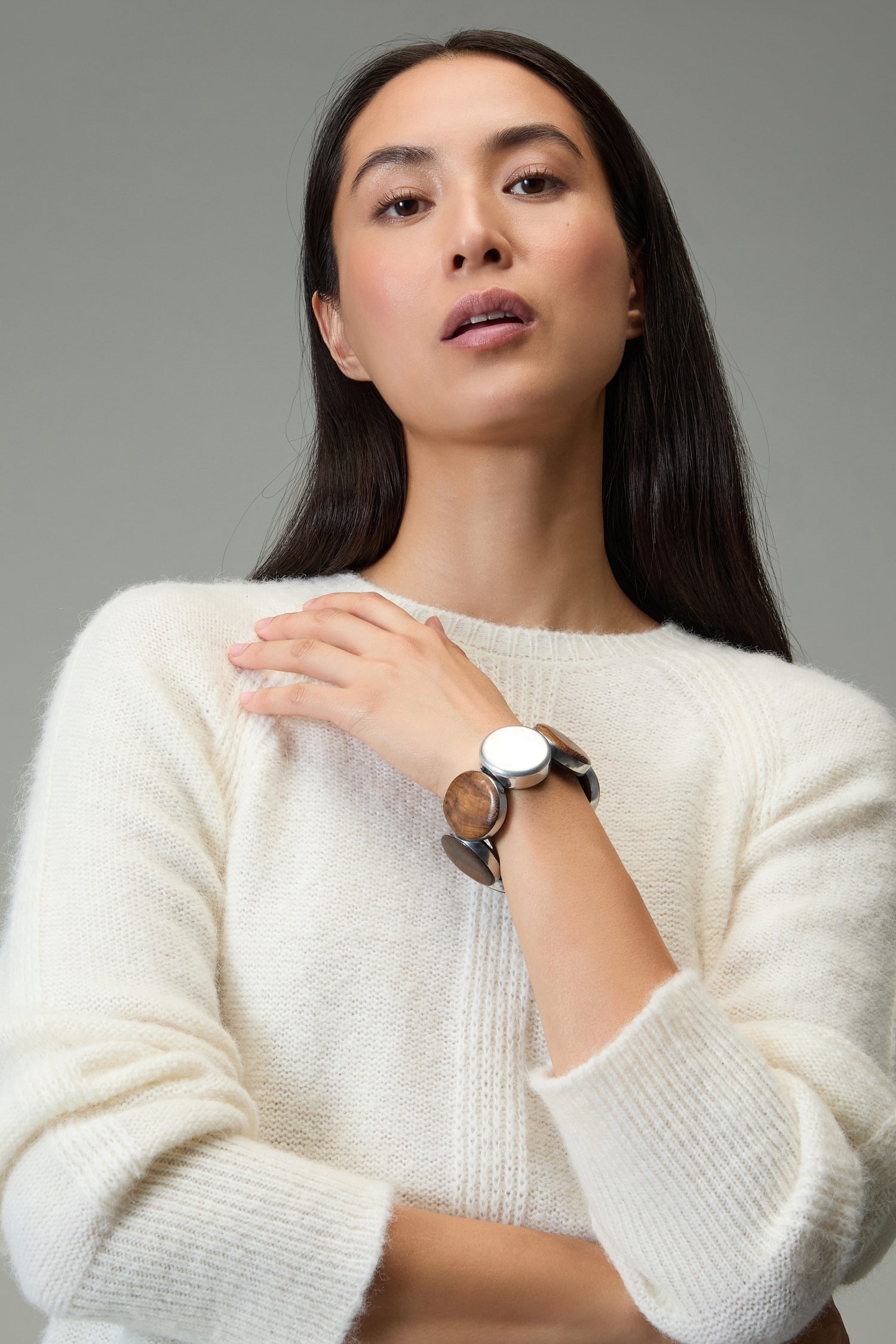 A woman with long dark hair wears a white sweater and a Silver Wooden Spheres Bracelet, posing with one hand on her shoulder against a plain gray background.