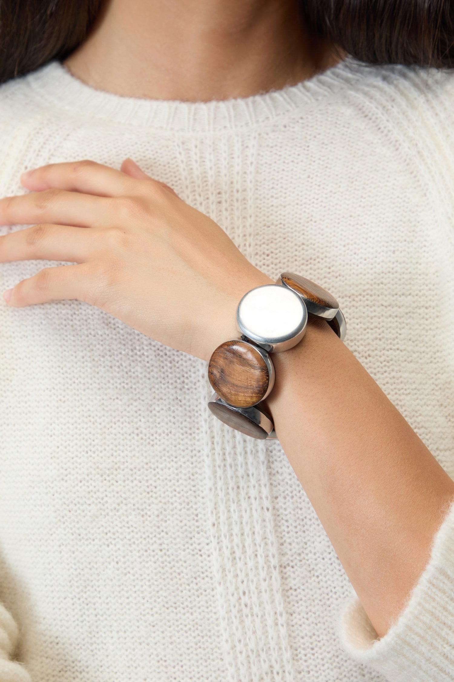 A person in a cream knit sweater showcases the Silver Wooden Spheres Bracelet—a handmade Spanish statement piece featuring large round wooden and metallic segments—on their wrist.
