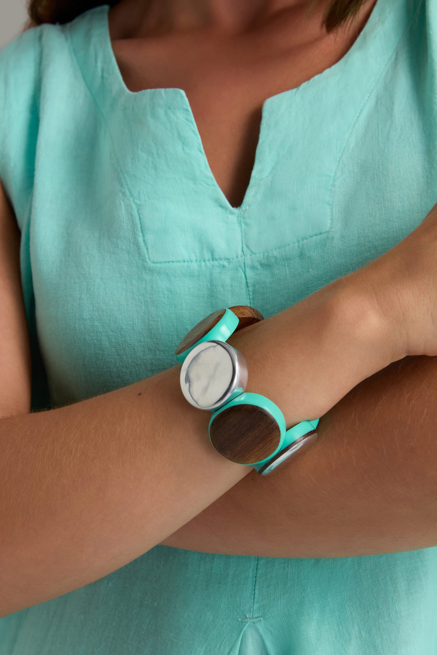 A person in a turquoise dress crosses their arms, showcasing the Wooden Spheres Bracelet, which features wood, silver, and turquoise elements along with circular white and patterned accents.