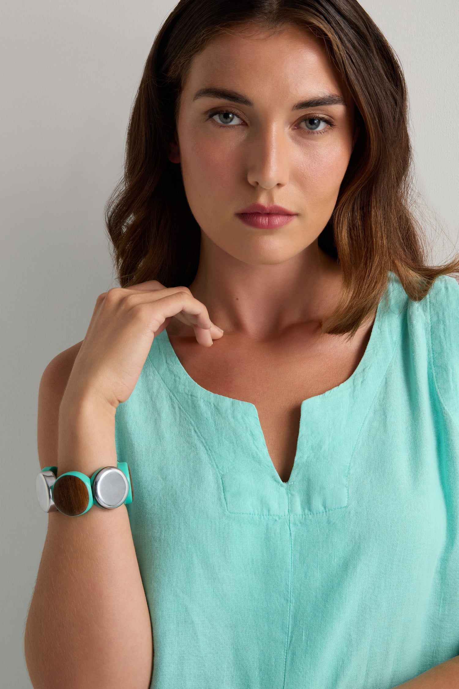 A woman in a sleeveless light turquoise top looks at the camera, wearing the Wooden Spheres Bracelet—featuring chunky wood, silver, and turquoise round elements—on her left wrist.