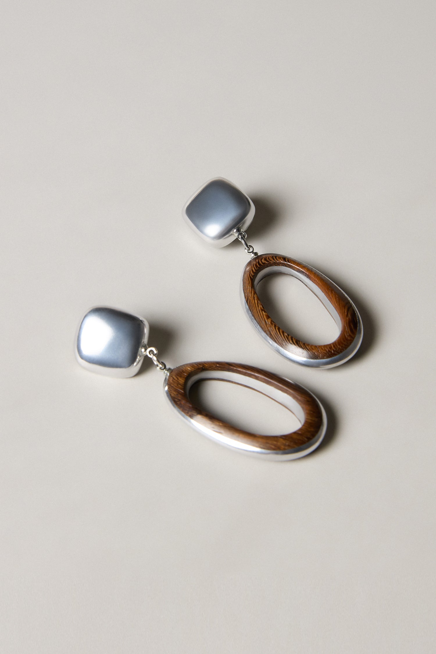 Handcrafted in Spain, the Wooden Silver Clip-On Earrings feature sculptural square silver studs and large oval wooden hoops, displayed on a neutral background.