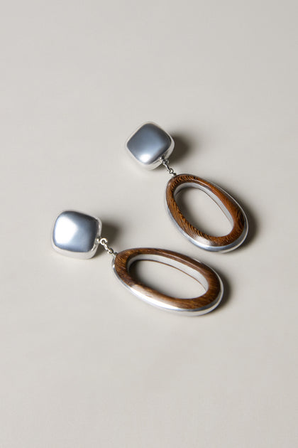 Handcrafted in Spain, the Wooden Silver Clip-On Earrings feature sculptural square silver studs and large oval wooden hoops, displayed on a neutral background.