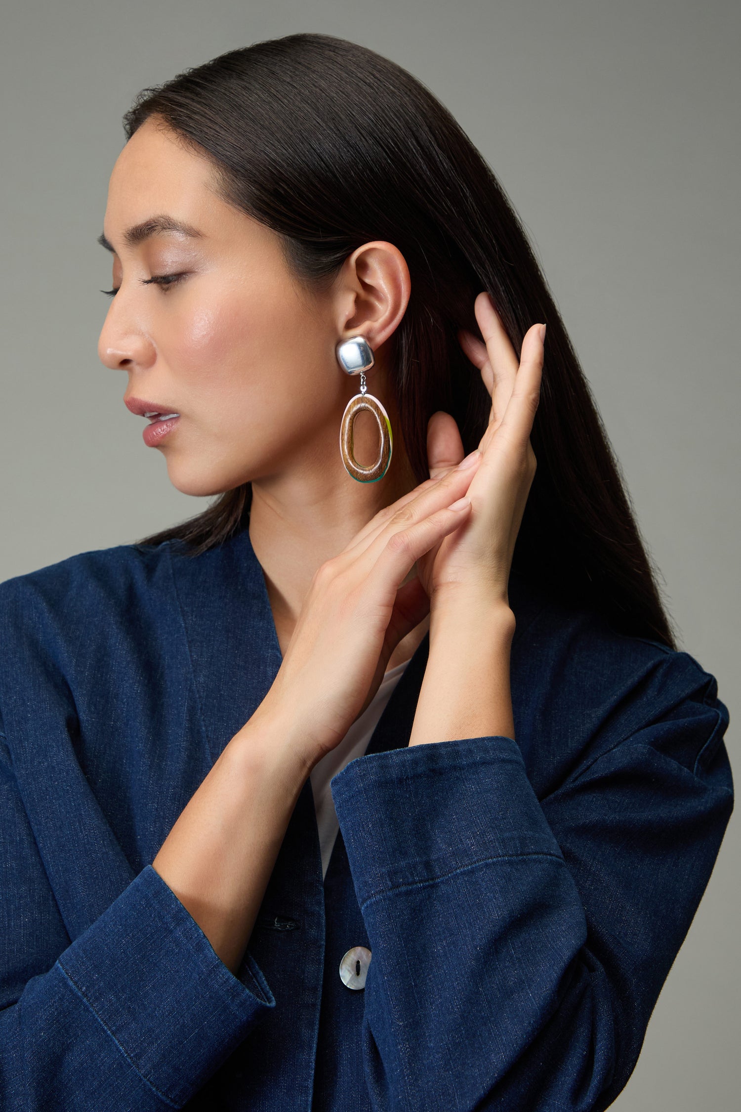A woman with straight dark hair wears a blue jacket and Rainbow Edge Wooden Clip-On Earrings, raising one hand near her ear and looking to the side against a neutral background.