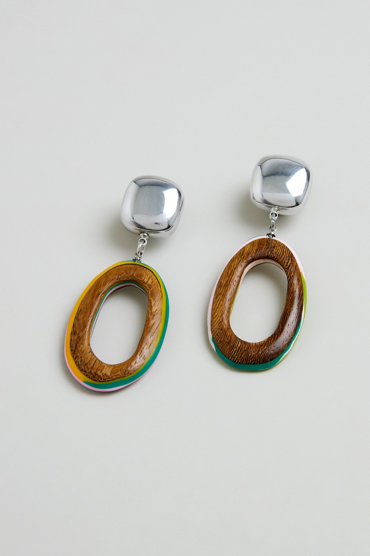 Introducing the Rainbow Edge Wooden Clip-On Earrings: handmade in Spain, these statement pieces feature chunky silver studs and oval wooden pendants with a subtle green accent on a neutral background.
