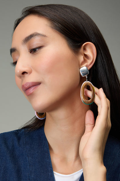 A woman with straight dark hair wears a blue top and Rainbow Edge Wooden Clip-On Earrings, gazing downward while holding one earring with her hand.