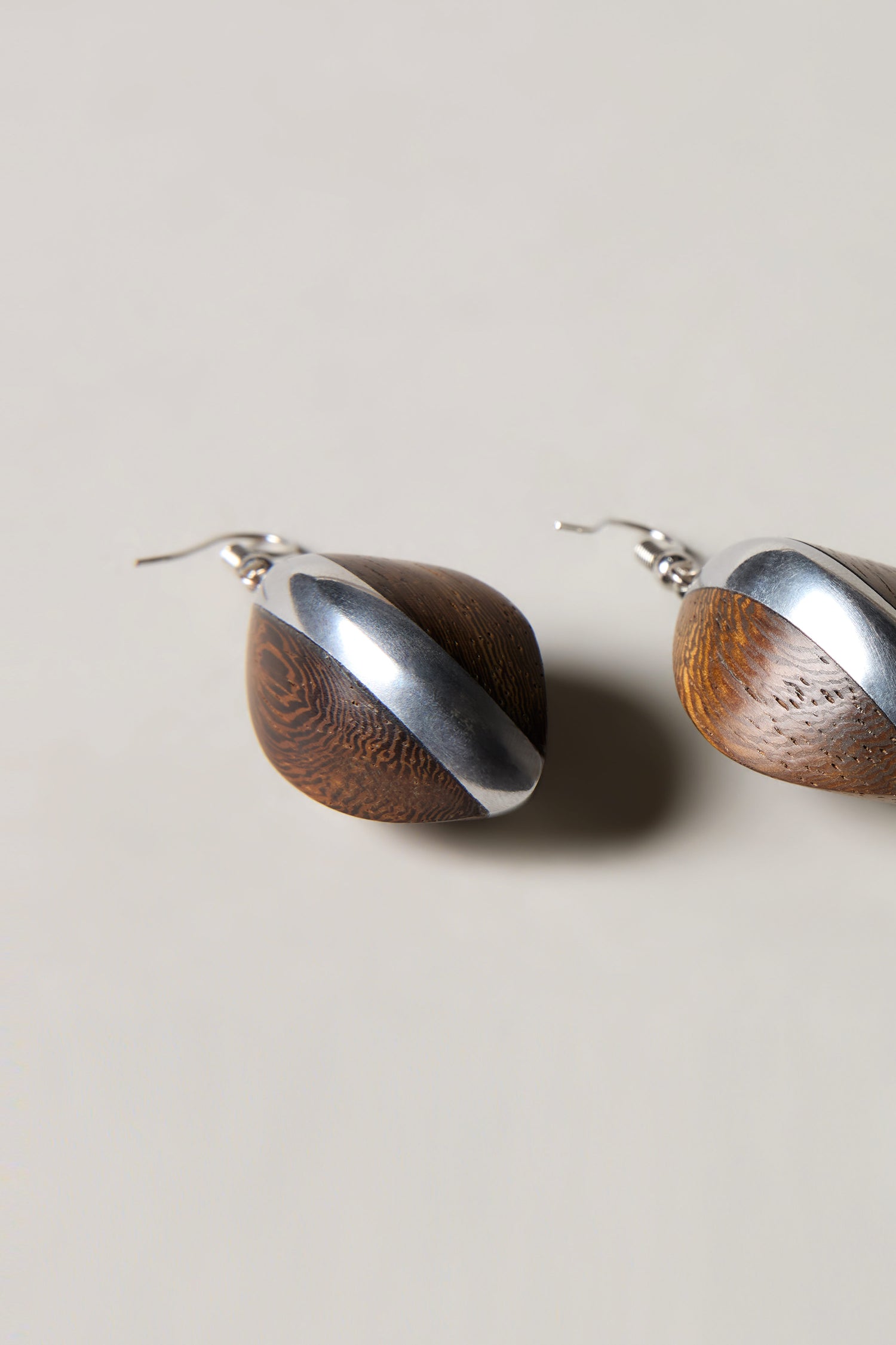 The Wooden Oval Pebble Earrings showcase rounded wooden shapes accented with diagonal silver metal, elegantly presented against a light background.