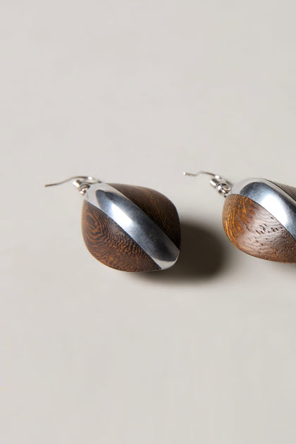 The Wooden Oval Pebble Earrings showcase rounded wooden shapes accented with diagonal silver metal, elegantly presented against a light background.