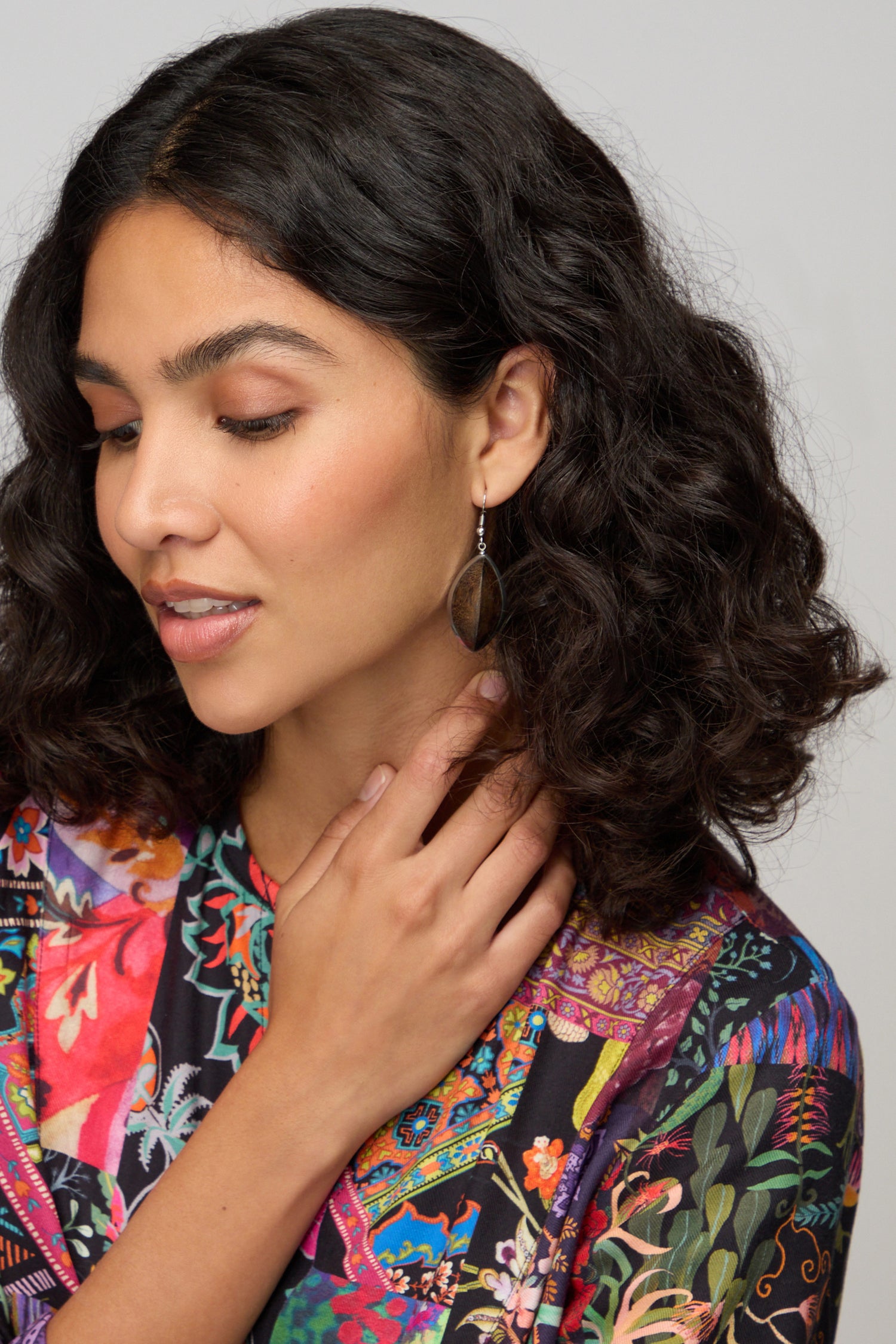 A woman with curly dark hair, wearing a colorful patterned top and Wooden Oval Pebble Earrings, looks down with her hand gently touching her neck.