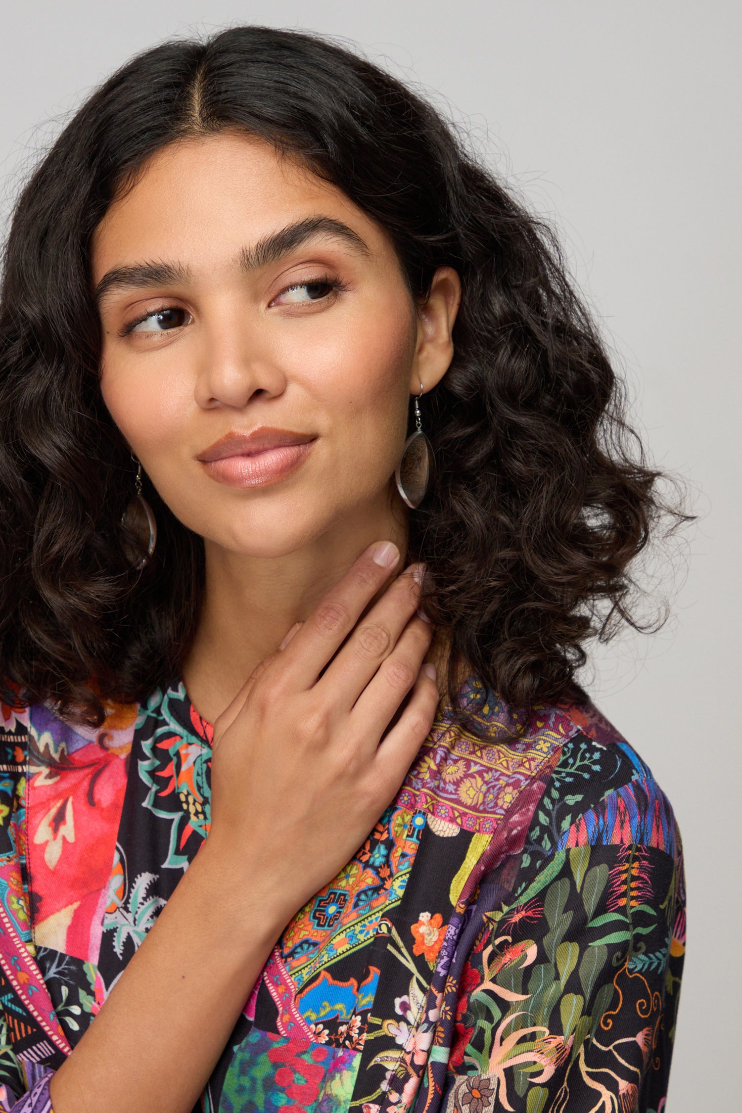 A woman with curly dark hair wears Wooden Oval Pebble Earrings and a colorful patterned top, looking to the side as she touches her neck with one hand.