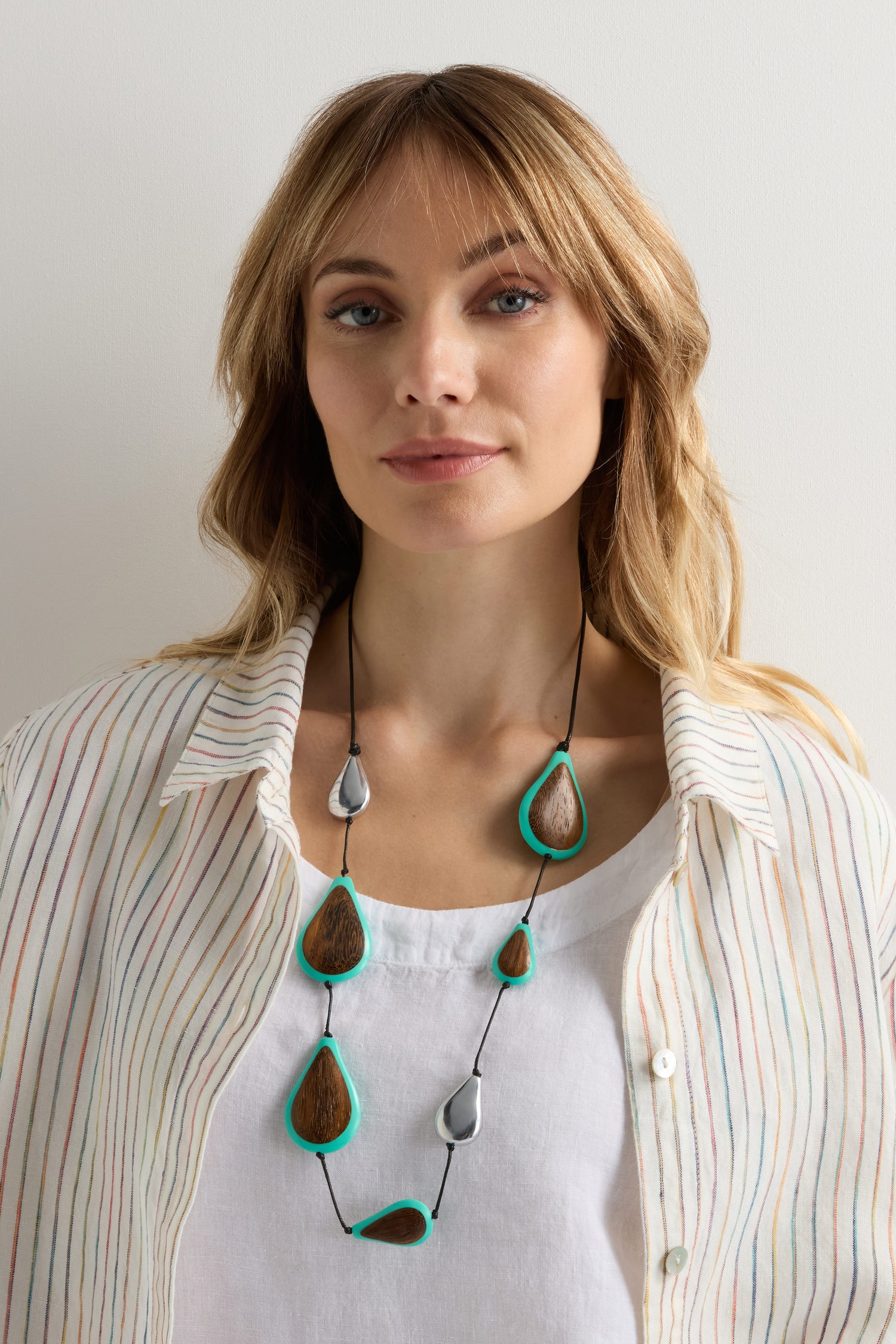 A woman with blonde hair wears a white shirt with pastel stripes and a white top underneath, accessorized with the Turquoise Teardrop Necklace.