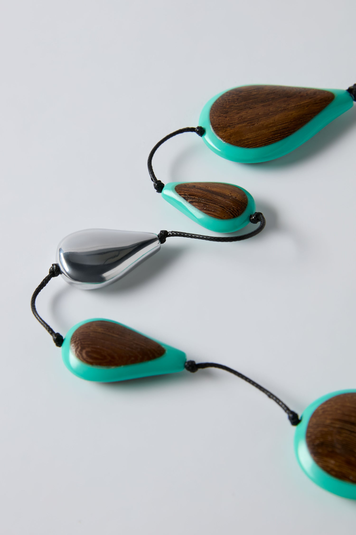 The Turquoise Teardrop Necklace features green-edged wooden beads with a single metallic silver teardrop, all strung on a black cord—an elegant piece by Ernesto de Barcelona.