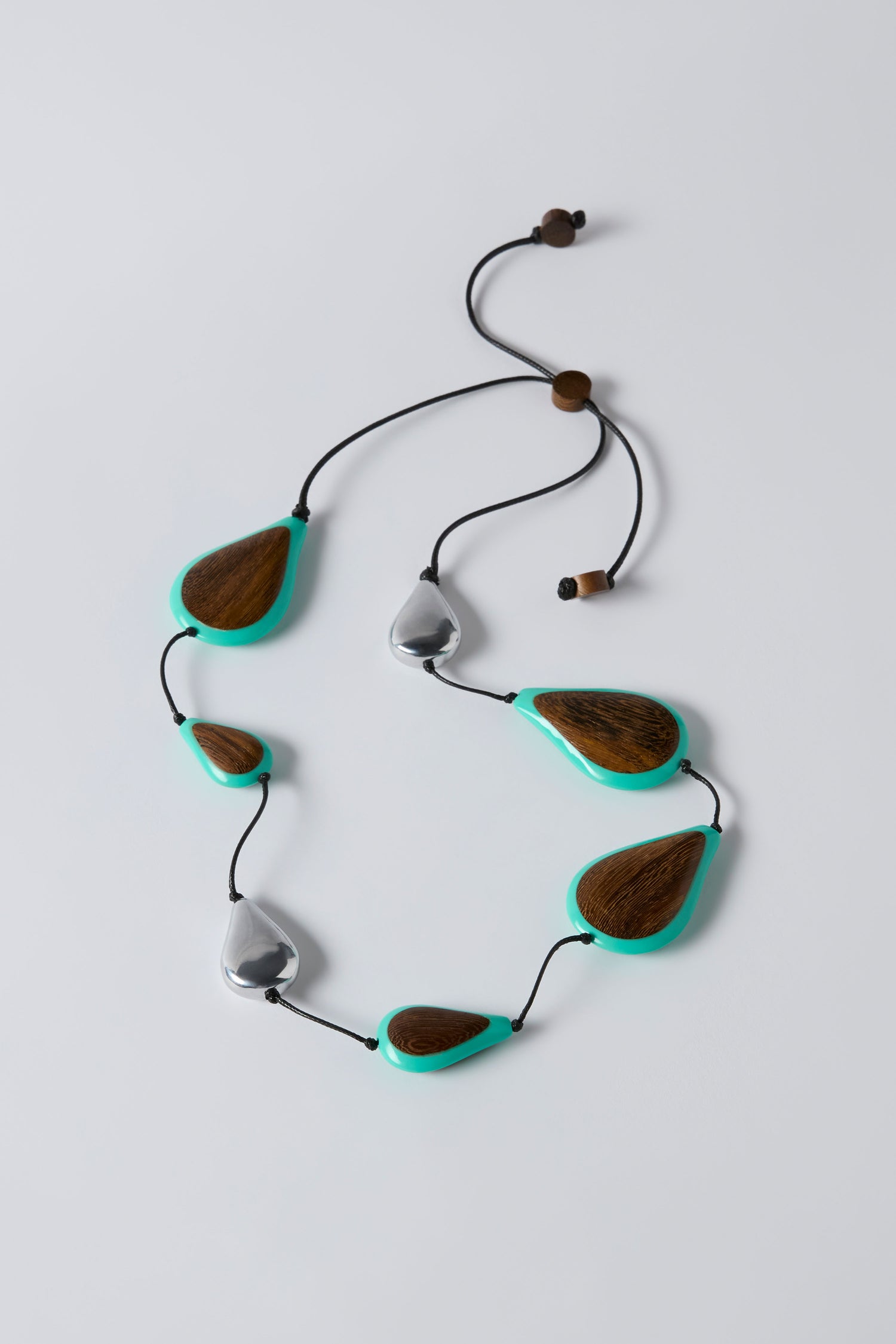The Turquoise Teardrop Necklace features an adjustable black cord, wooden and silver-tone beads, and turquoise accents, displayed on a plain light background.