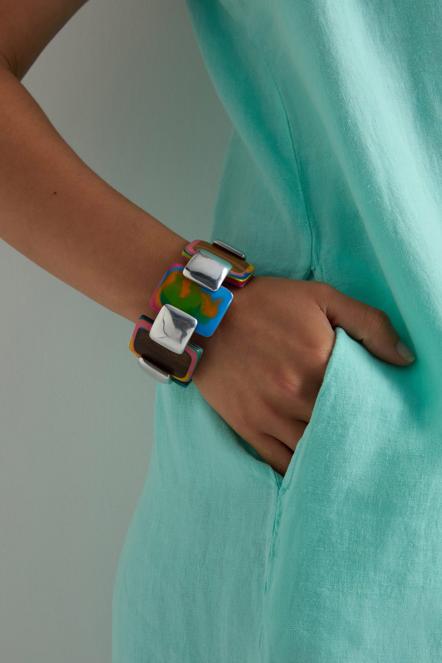 A person wearing a turquoise garment with their hand in their pocket showcases the Colourburst Squares Links Bracelet, a chunky handcrafted piece made of marbled resin, on their wrist.