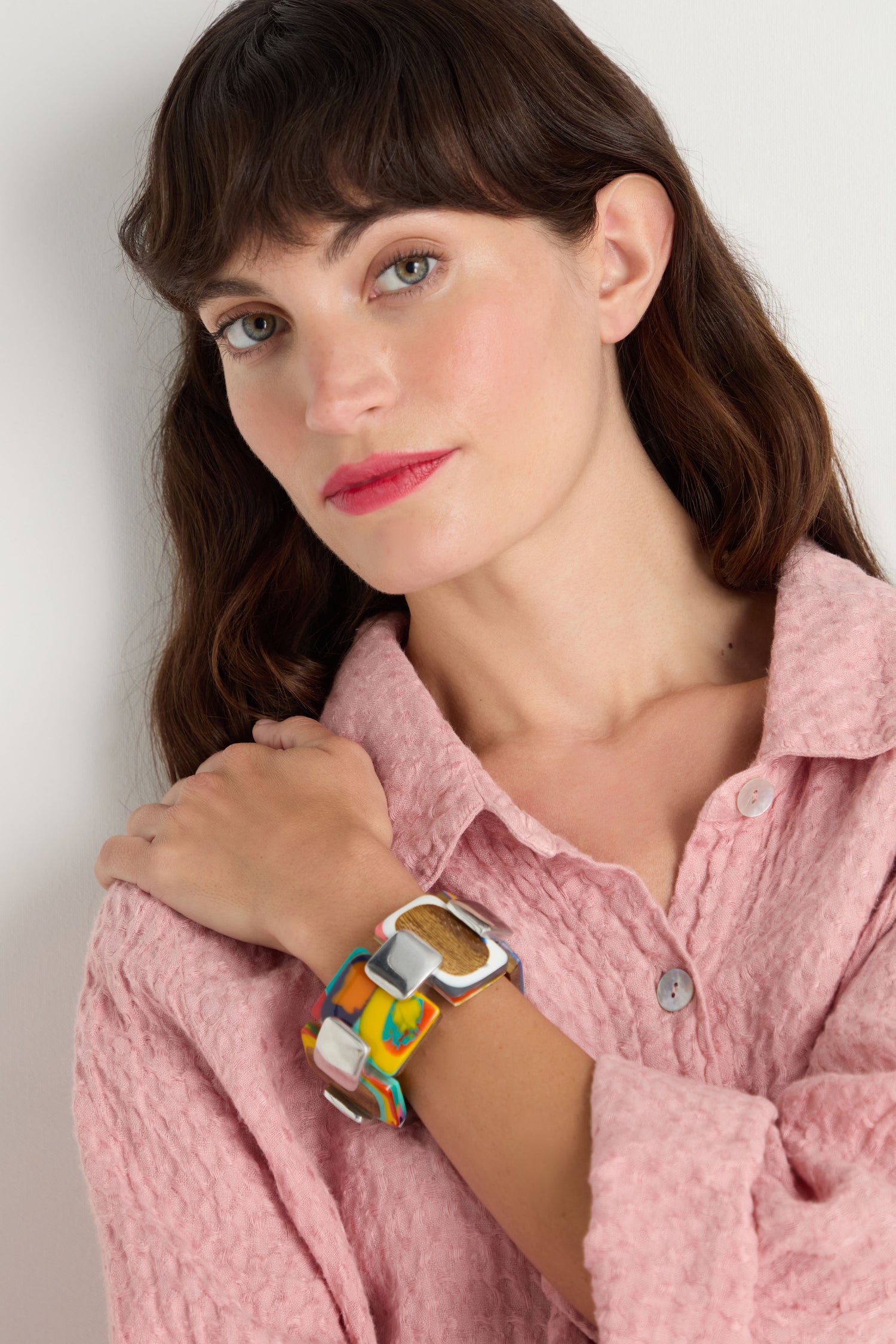 A woman wearing a textured pink button-up shirt and the Colourburst Squares Link Bracelet poses with her hand on her shoulder, looking at the camera.