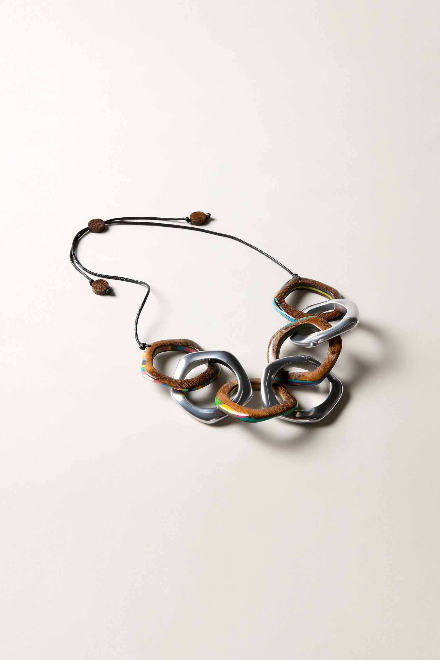 The Olympus Links Necklace is a chunky handmade piece with large marbled resin interlocking links in brown, gray, and metallic tones, finished with an adjustable black cord and wooden bead accents.