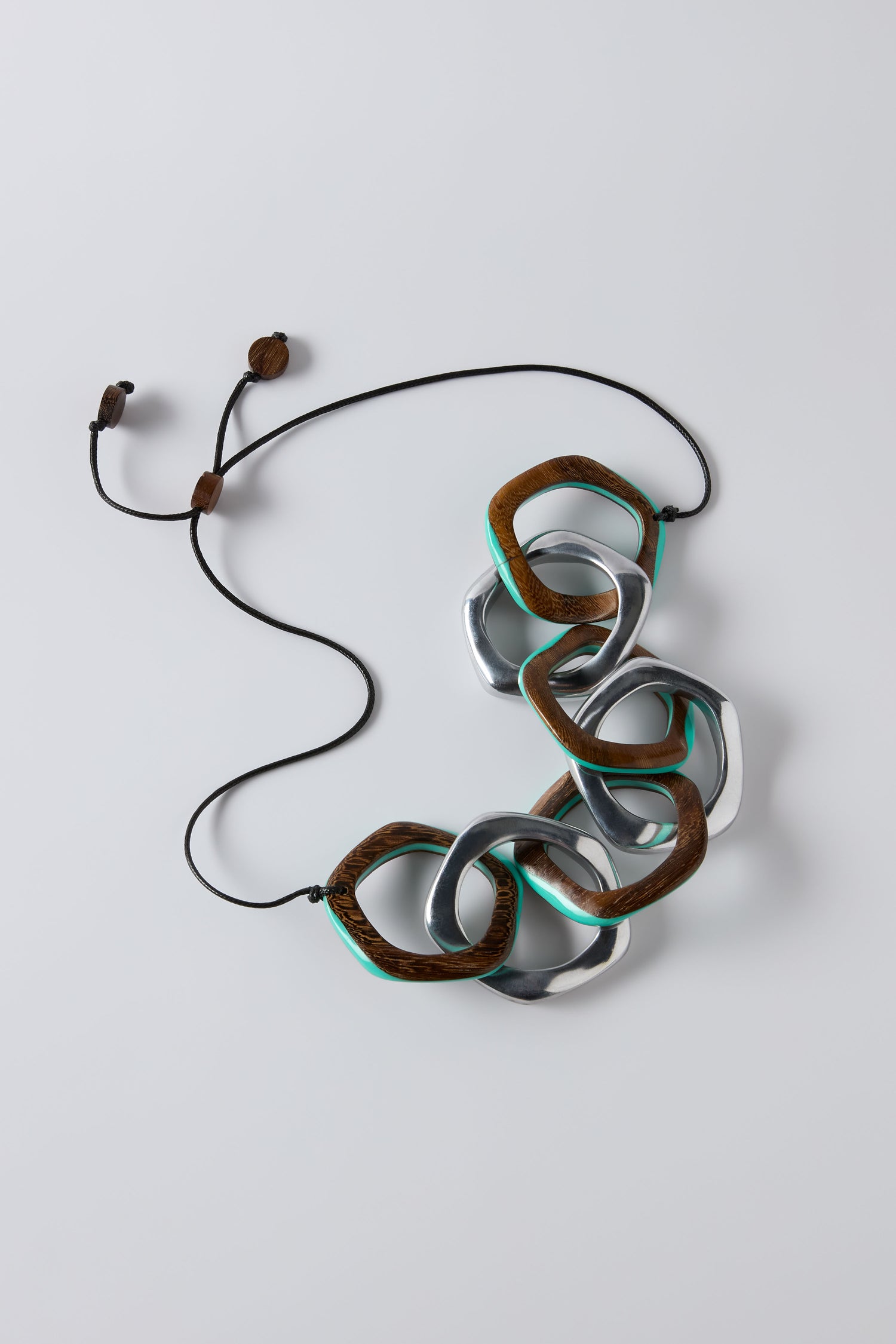 Make a statement with the Turquoise Olympus Links Necklace—oversized interlocking metallic and wood-textured links with an adjustable black cord and wooden bead ends, showcased on a light background.