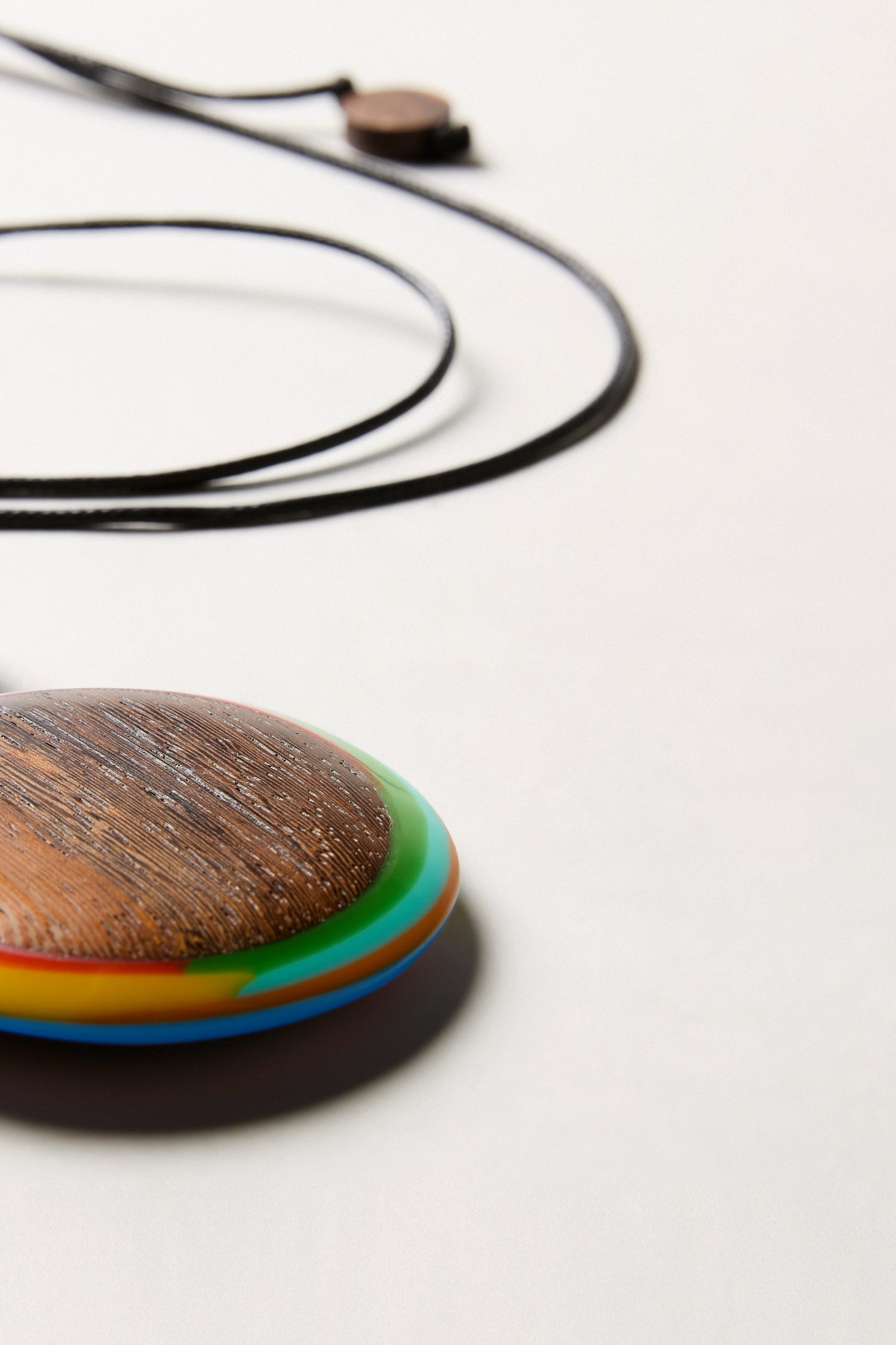 A close-up of the Colourburst Double Sphere Pendant, a round wooden pendant with a colorful edge on an adjustable cord necklace, shown on a light surface.