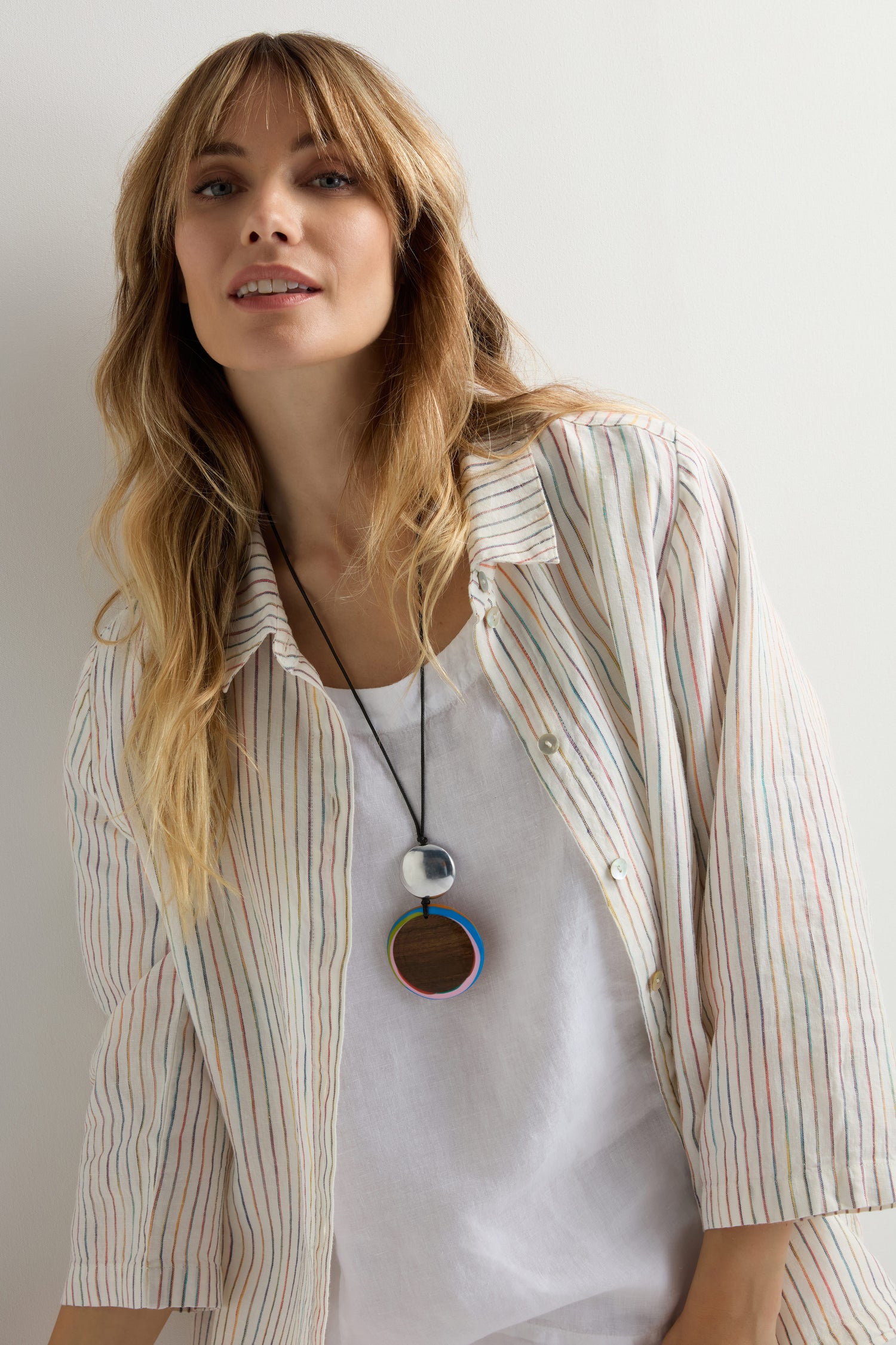 A woman with long blonde hair wears a white shirt with pastel stripes, a white top underneath, and the Colourburst Double Sphere Pendant, handmade in Barcelona, standing against a white background.