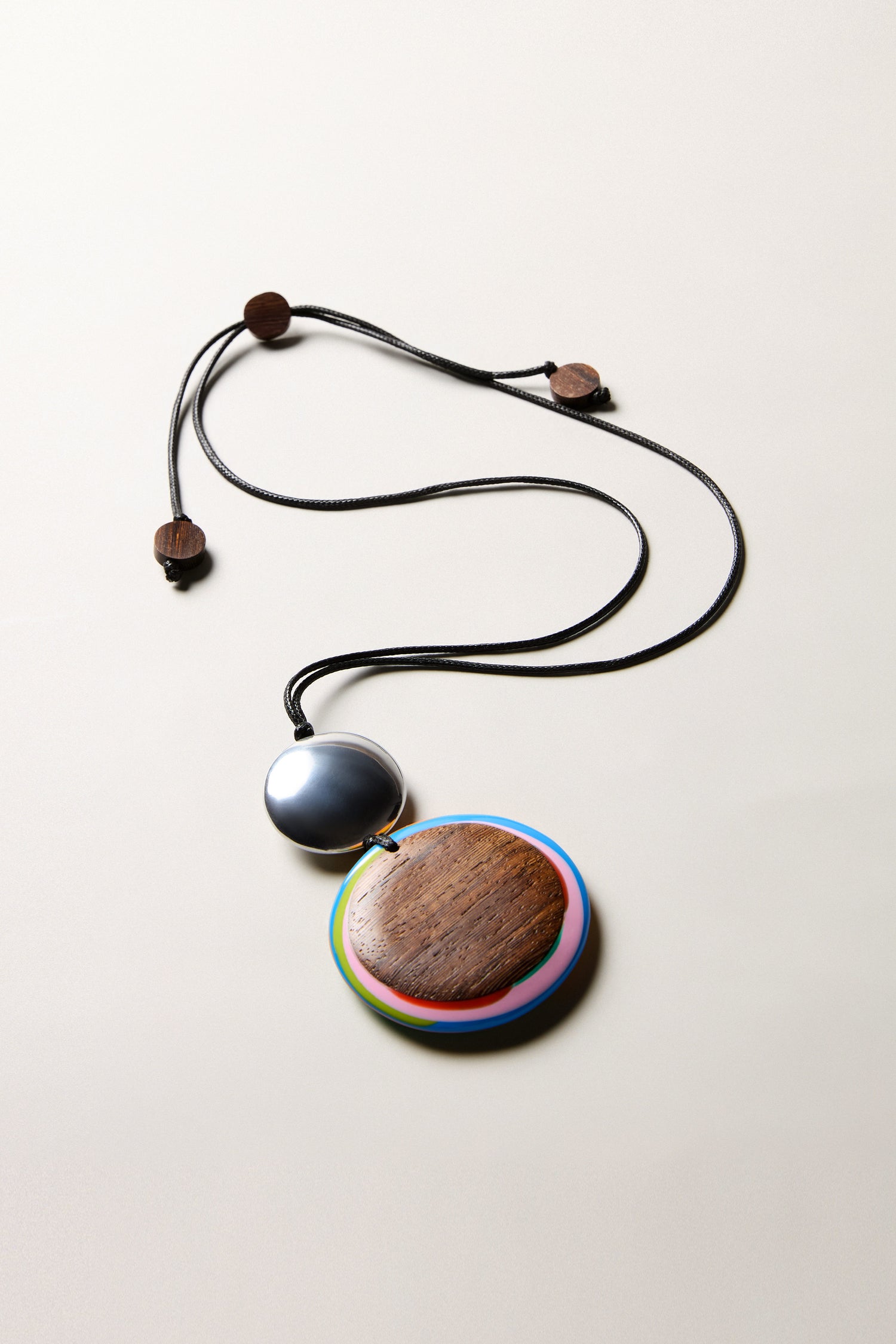 The Colourburst Double Sphere Pendant features two circular pendants—one metallic, one wood with a colorful border—on an adjustable cord with wooden bead clasps. Handmade in Barcelona, it's shown on a light background.