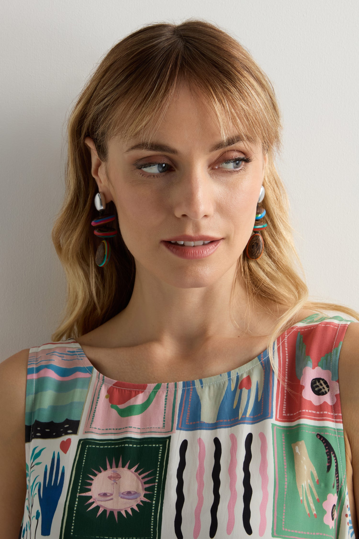 A blonde woman wearing Colourburst Wooden Pebble Earrings and a sleeveless, multicolored artistic top looks slightly to her left.