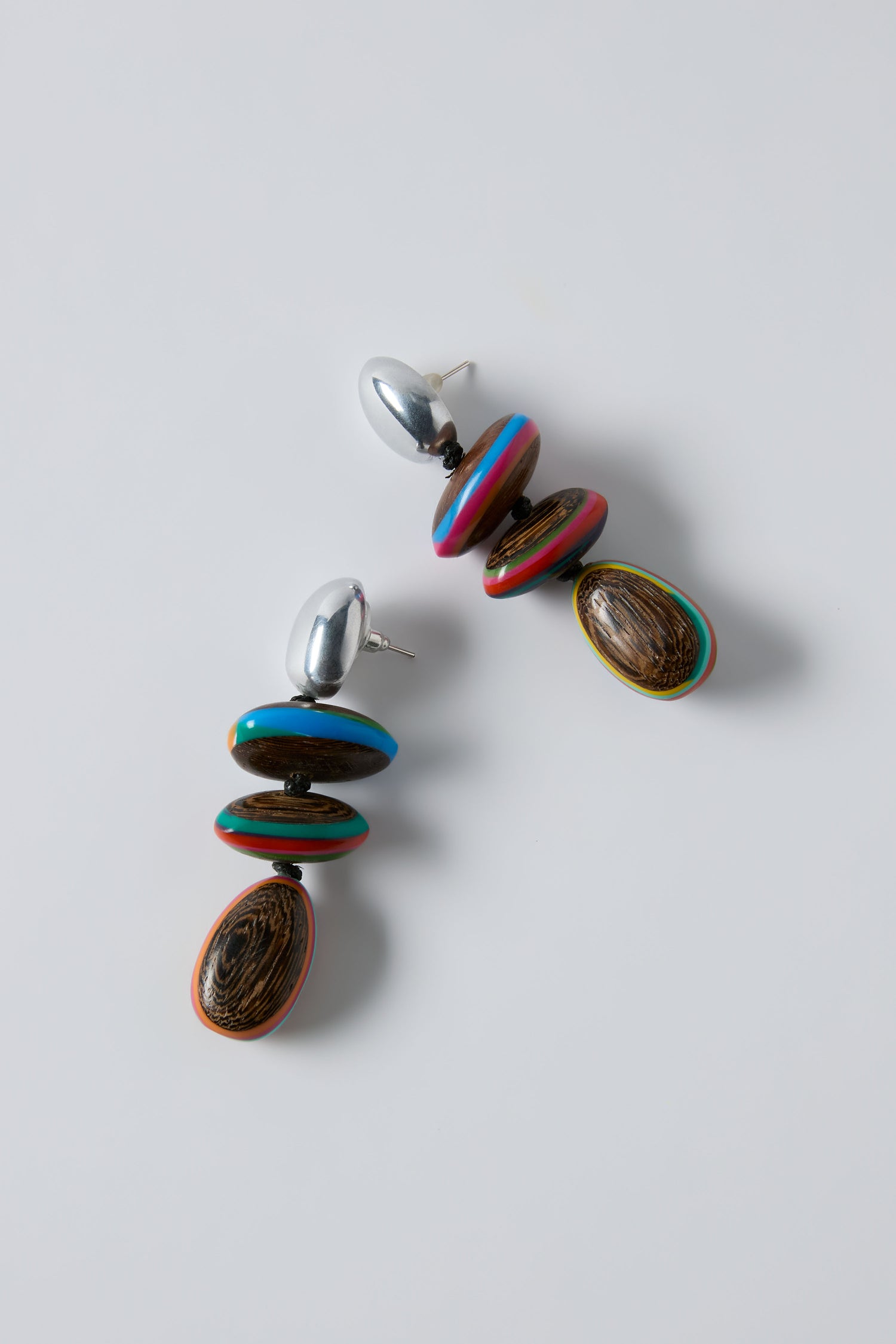 Colourburst Wooden Pebble Earrings by Ernesto de Barcelona feature silver studs, stacked colorful beads, and oval wood grain pendants, shown on a white background.