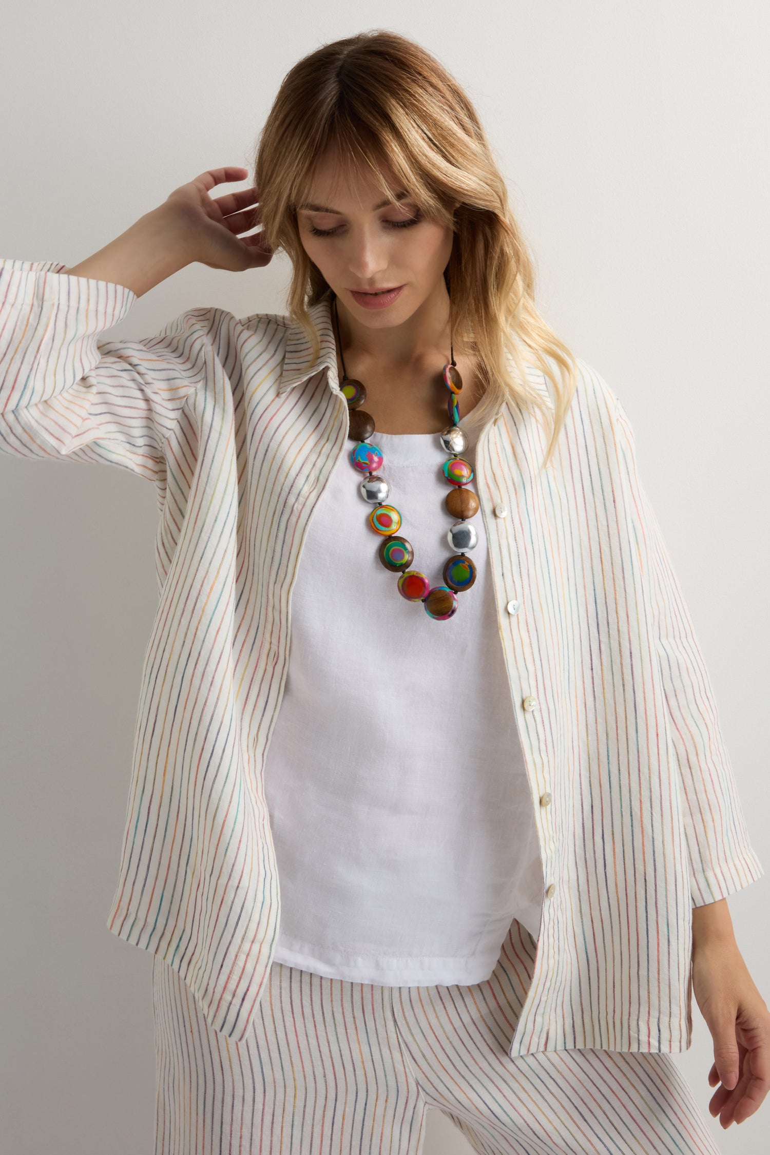 A woman in a loose, striped button-up shirt over a white top and matching pants poses against a light background, accessorized with the playful Rainbow Riverstone Spheres Necklace.