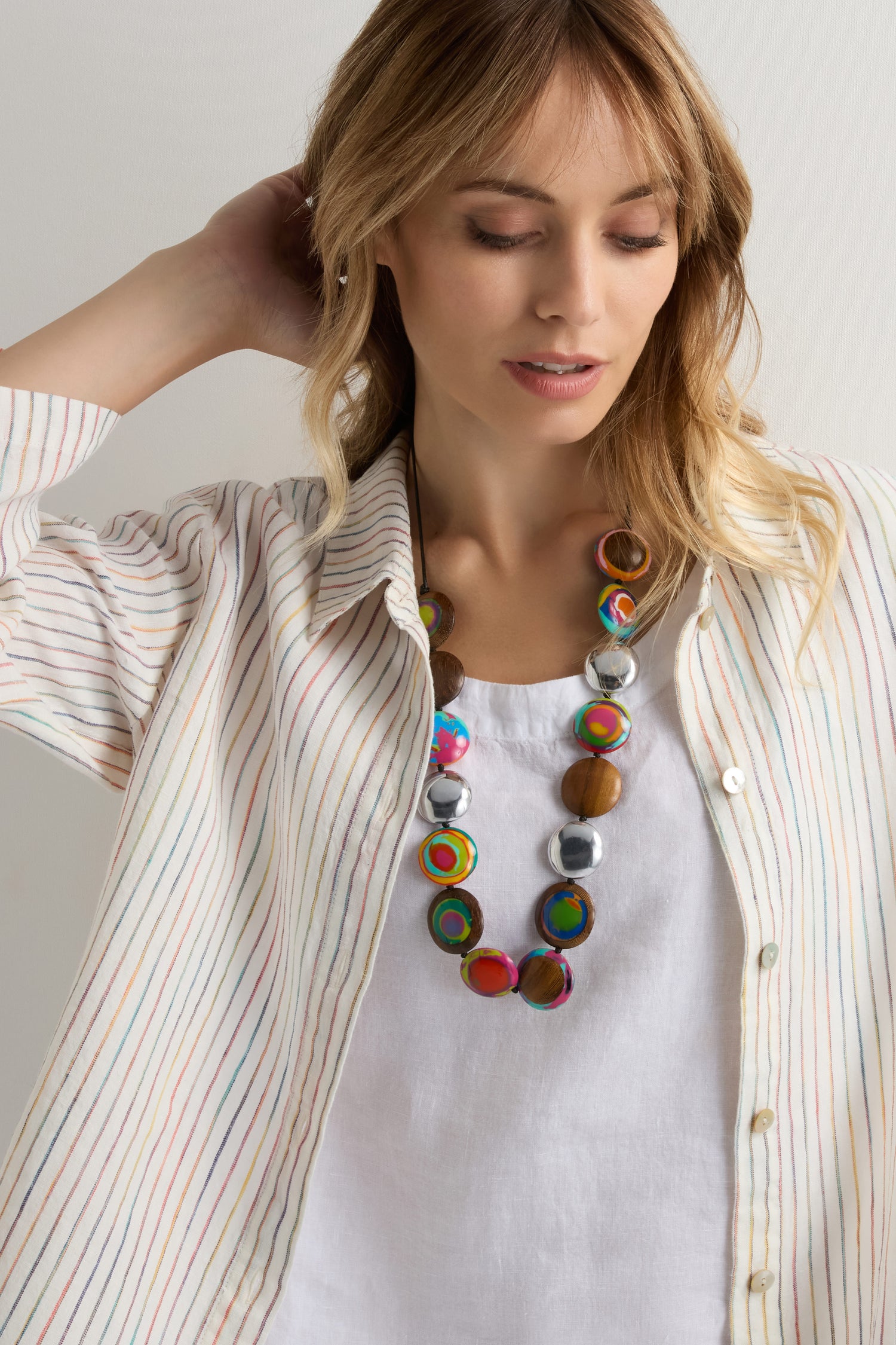 Woman wearing a striped button-up shirt over a white top, gazing down, with one hand behind her head and accessorized with the playful Rainbow Riverstone Spheres Necklace.