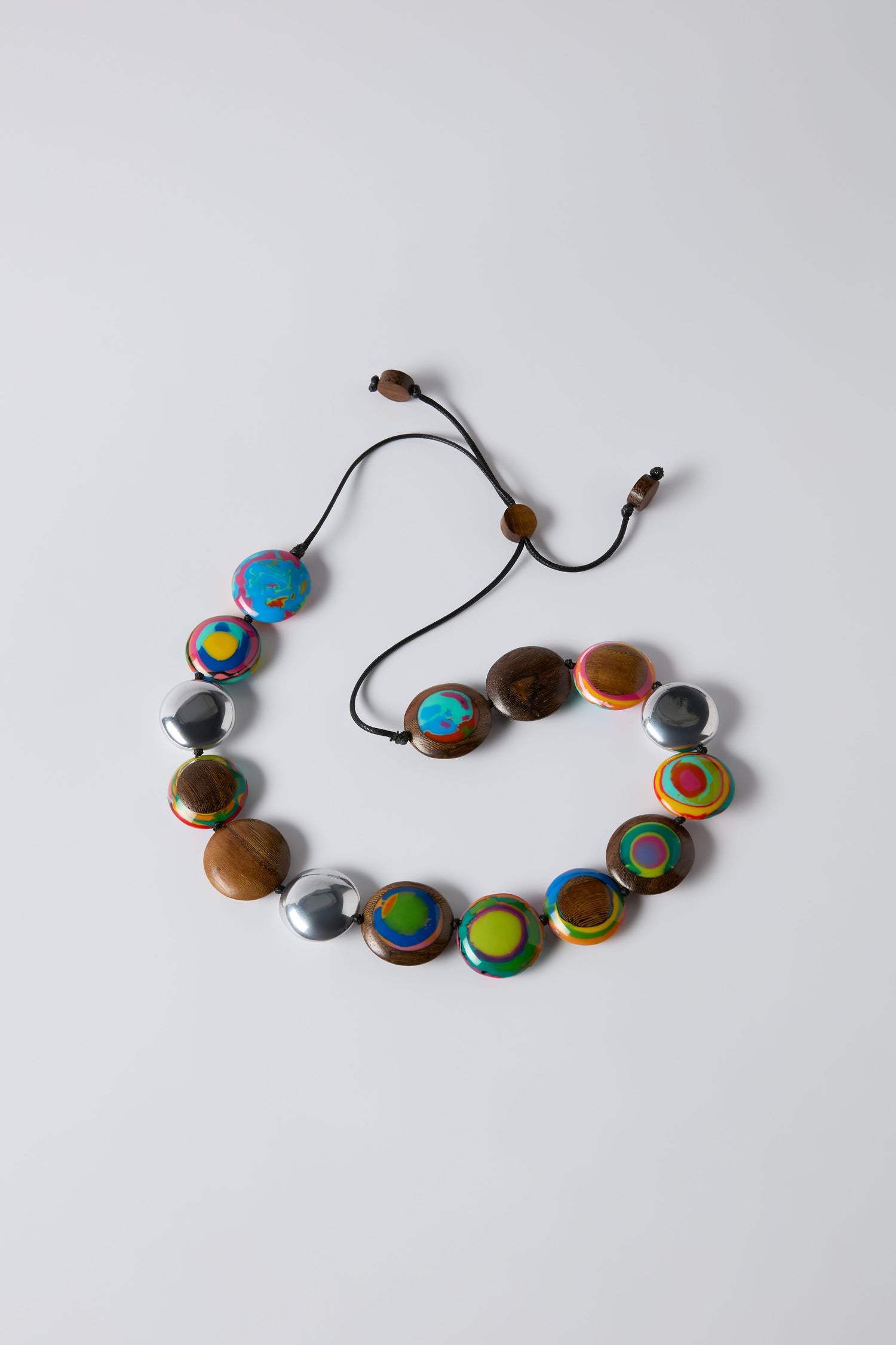 The Rainbow Riverstone Spheres Necklace showcases colorful round beads of wood, metal, and painted finishes on an adjustable black cord, presented against a plain white background.