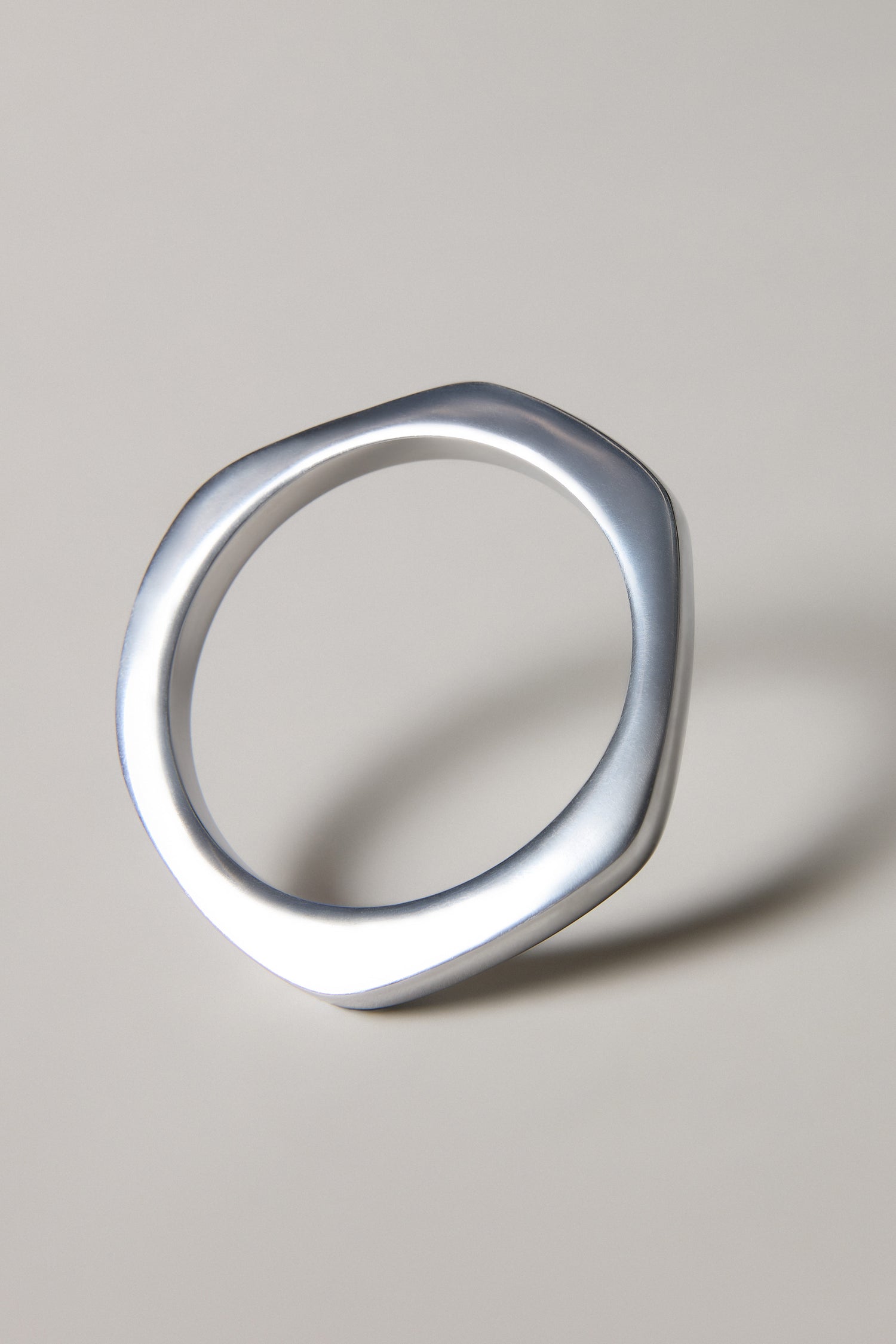 The Solar Silver Bangle is a handmade Spanish bracelet featuring a silver, geometric design with a slightly irregular, rounded polygon shape—an elegant accessory casting a subtle shadow on a neutral background.