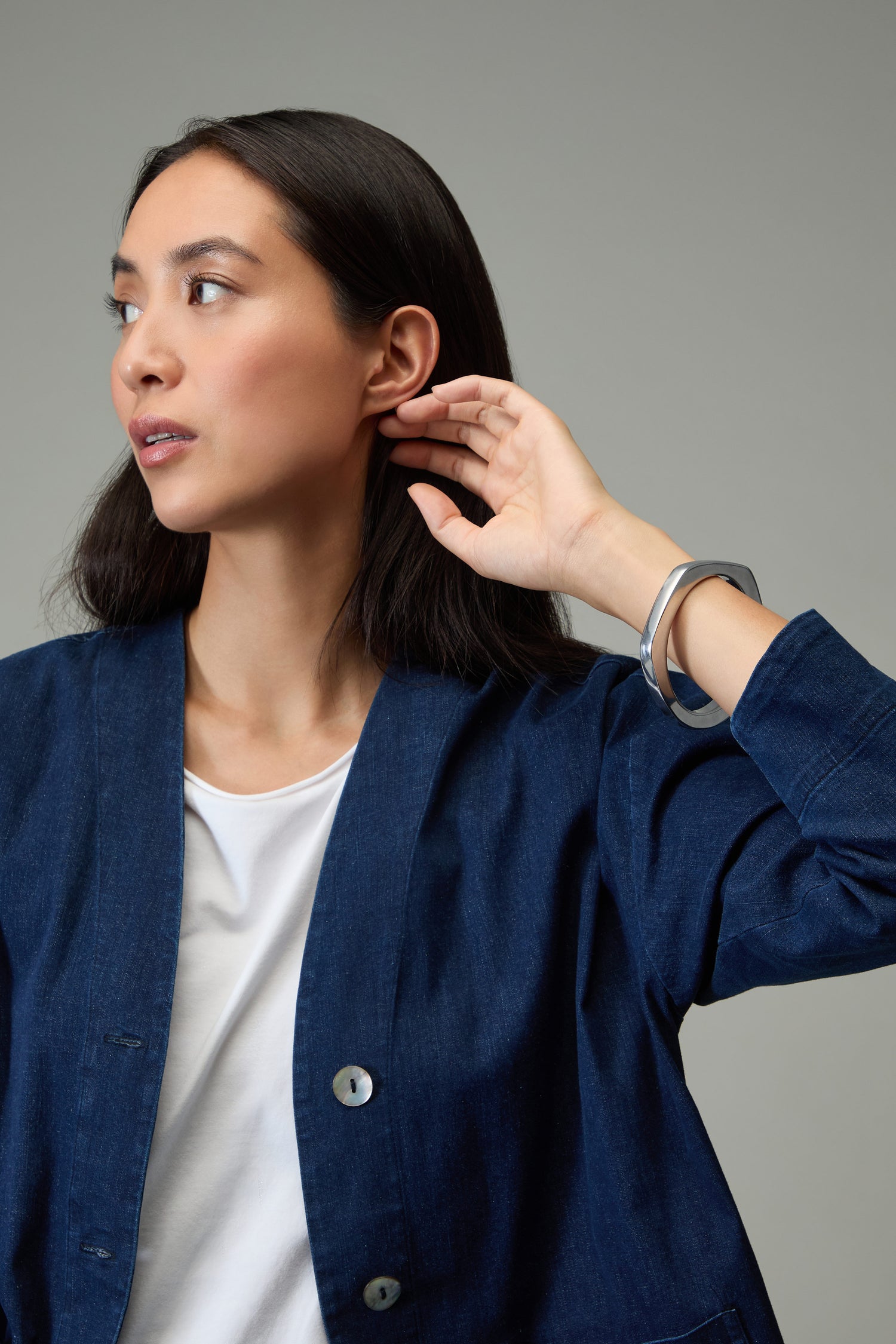 A woman in a blue jacket and white shirt looks to the side, touching her ear with one hand. She is wearing the Solar Silver Bangle, a striking piece inspired by handmade Spanish jewelry.