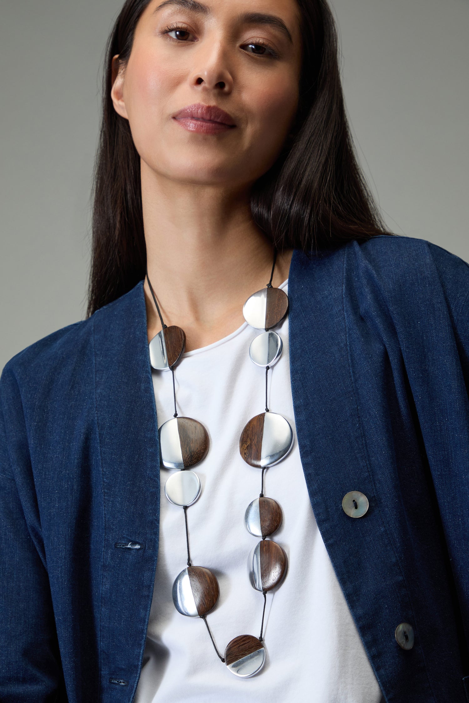 A woman in a blue jacket and white shirt wears the Solar Spheres Necklace, a statement piece featuring alternating round wood and silver elements, while standing against a neutral background.