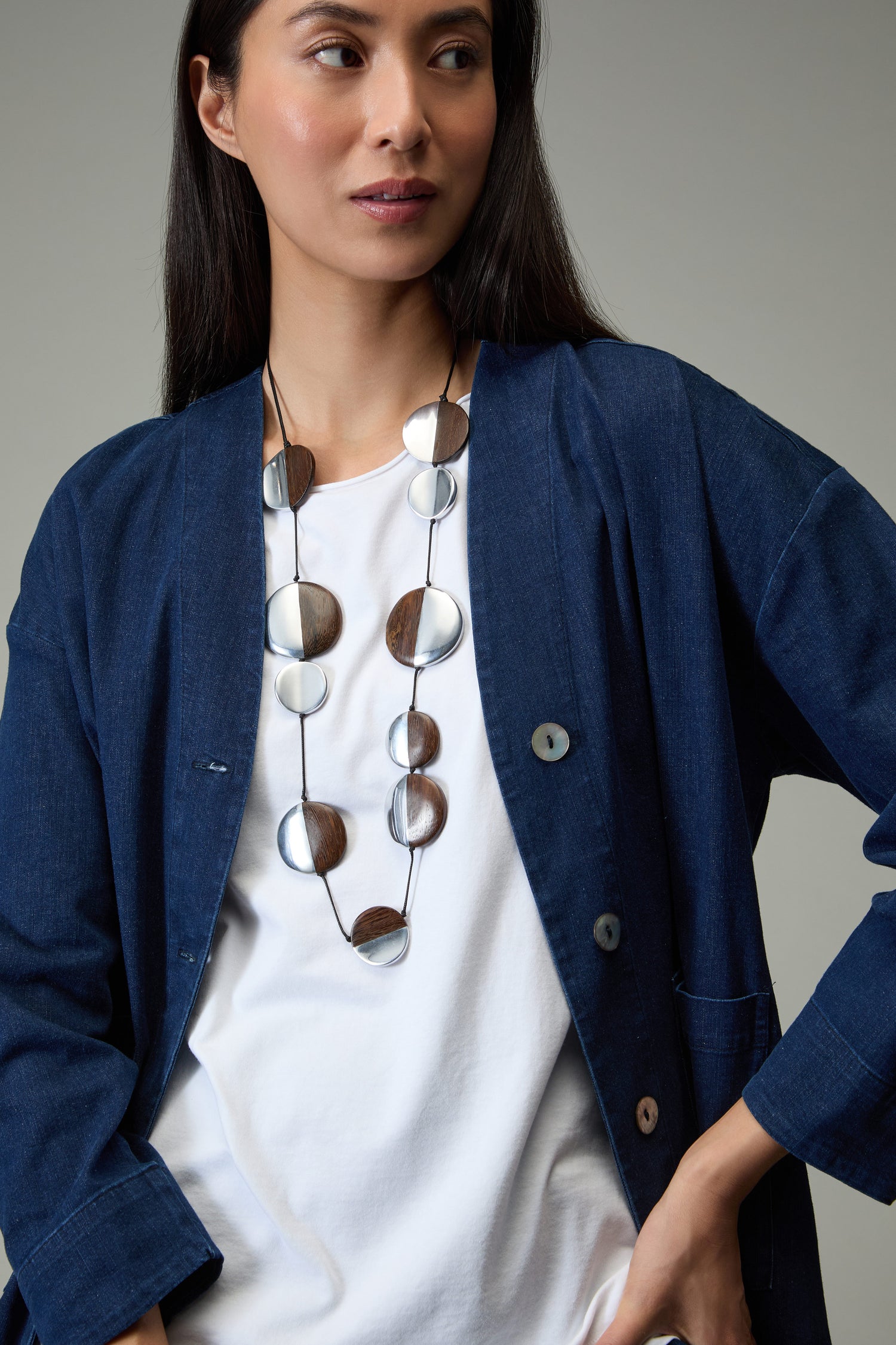 A woman in a blue button-up jacket over a white t-shirt wears the Solar Spheres Necklace—an eye-catching statement piece featuring large circular pendants—while standing against a neutral background.