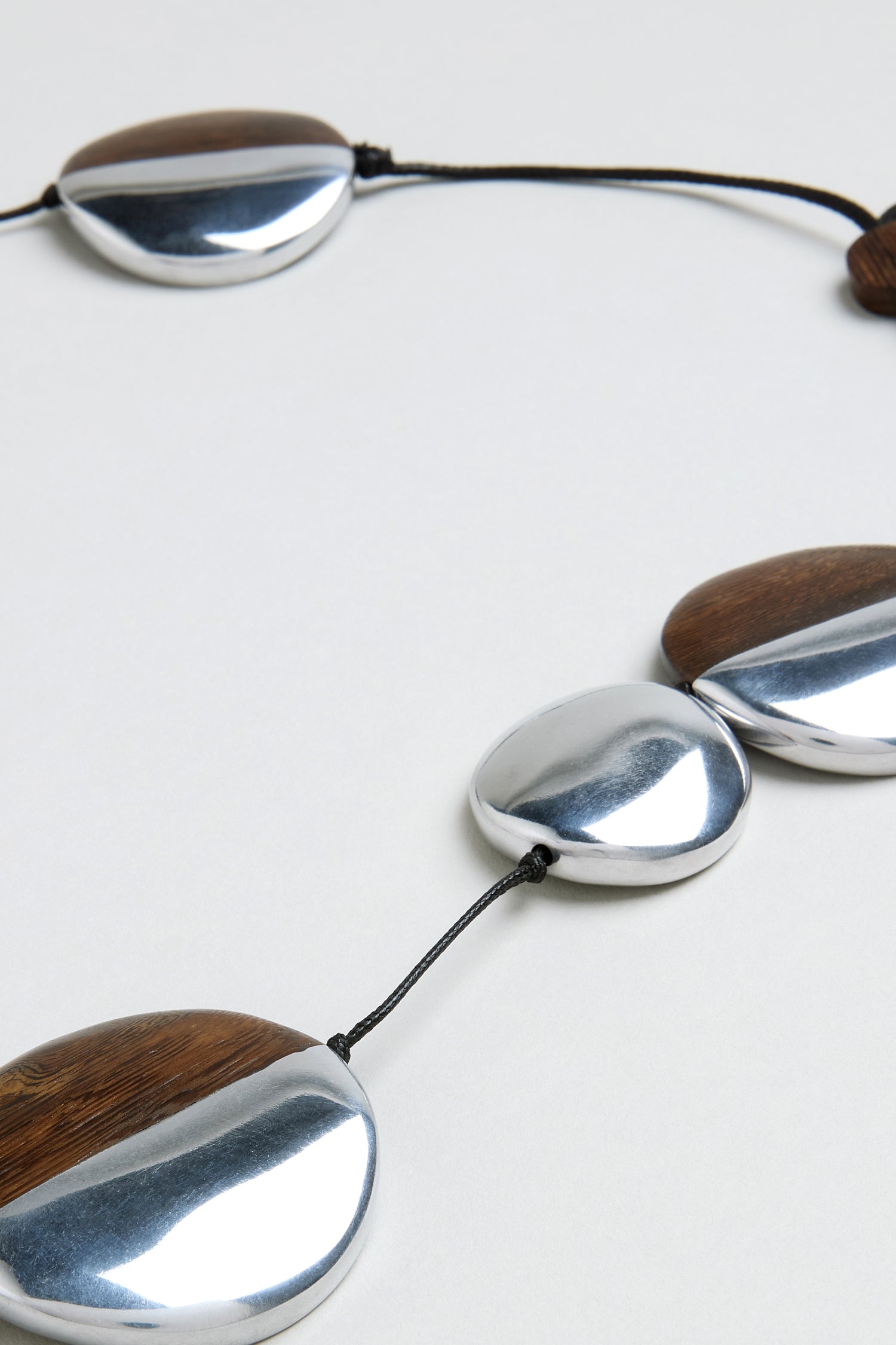 A close-up of the Solar Spheres Necklace, showcasing large, flat oval beads crafted from silver metal and dark wood, all strung on a black cord.