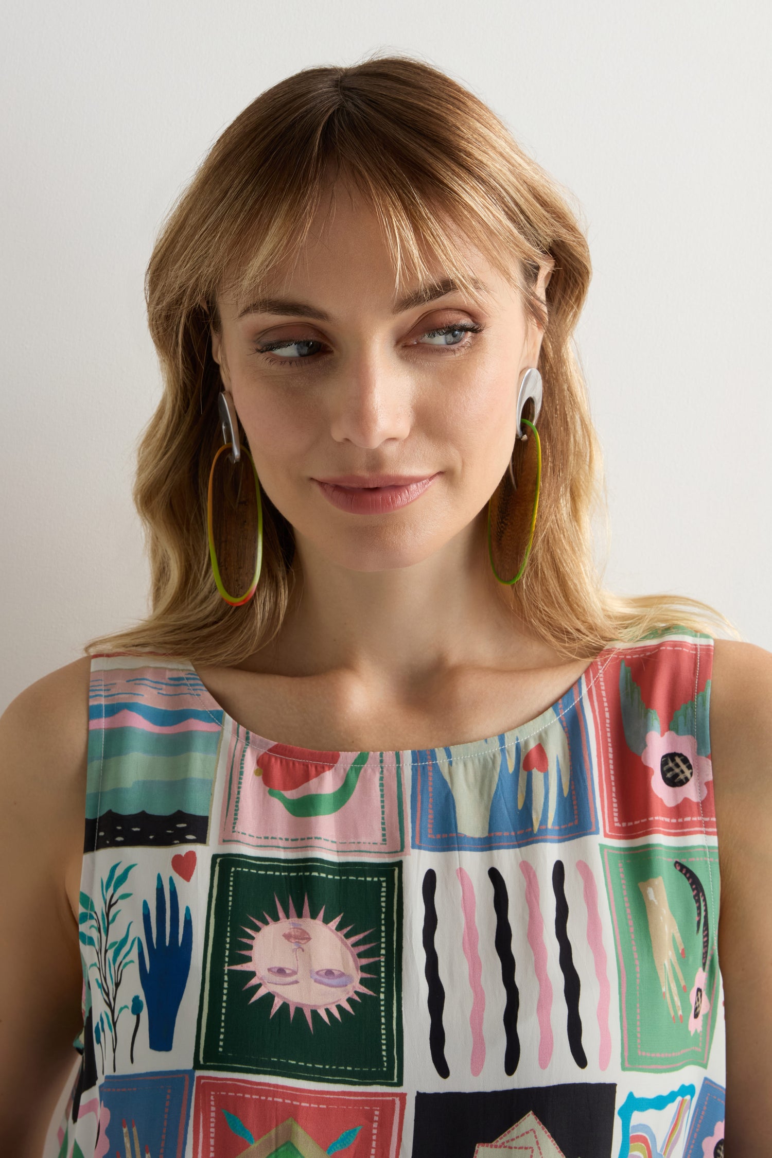 A woman with blonde hair wears Rainbow Wooden Oval Links Earrings and a sleeveless top with colorful, square abstract designs. She looks slightly to the side, smiling gently.