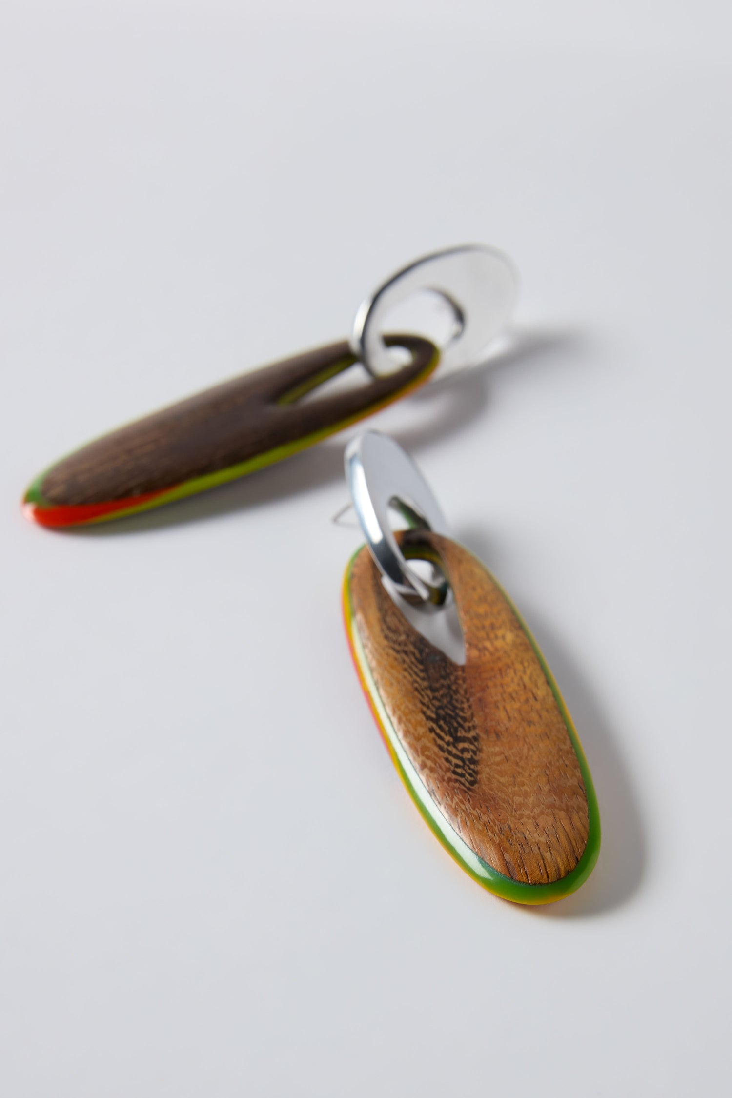 The Rainbow Wooden Oval Links Earrings by Ernesto de Barcelona feature sculptural oval drops with green and red-edged wooden pendants and silver metal loops, artfully displayed on a plain white surface.