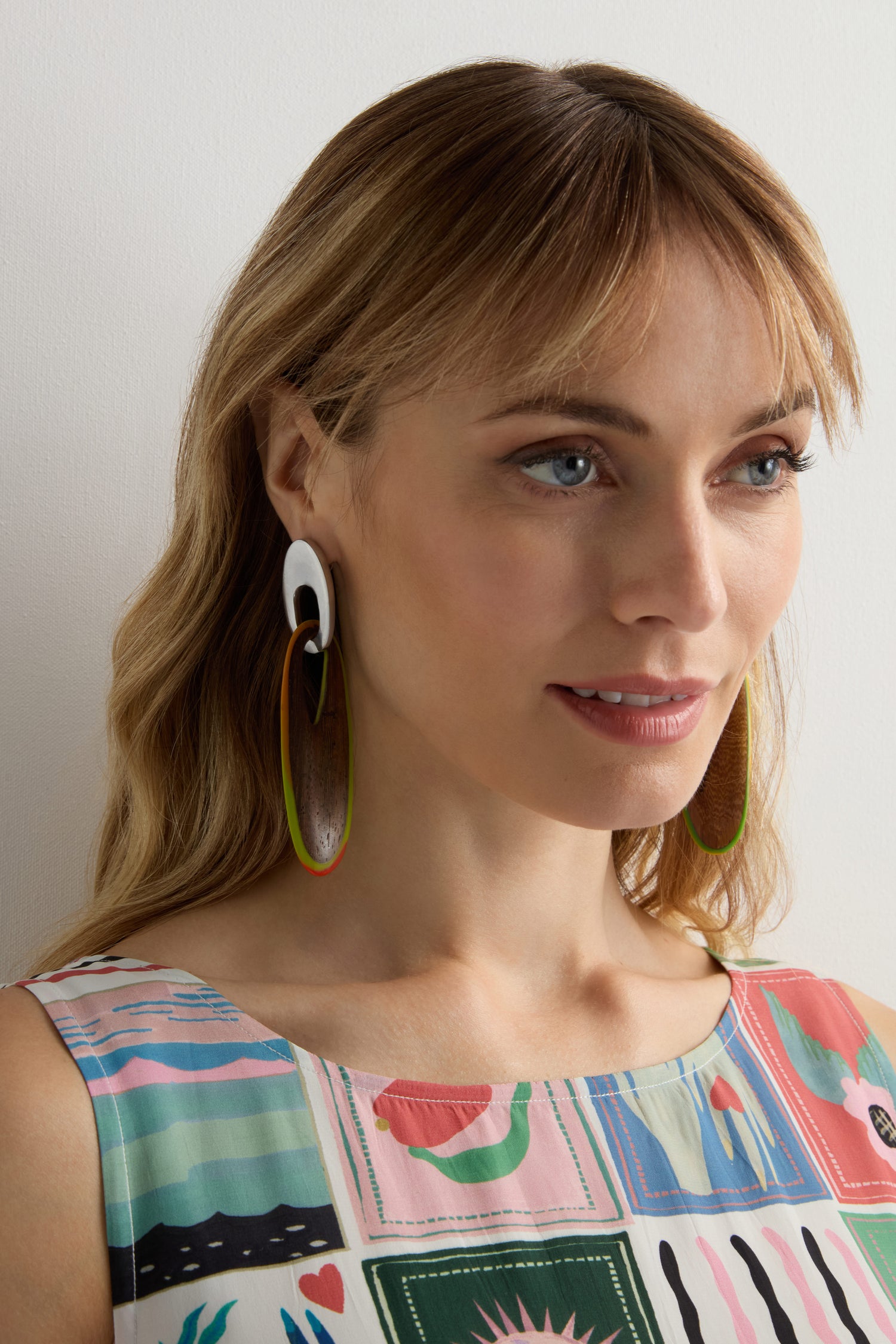 A woman with light brown hair wears Rainbow Wooden Oval Links Earrings and a sleeveless patterned top, standing against a plain white background.