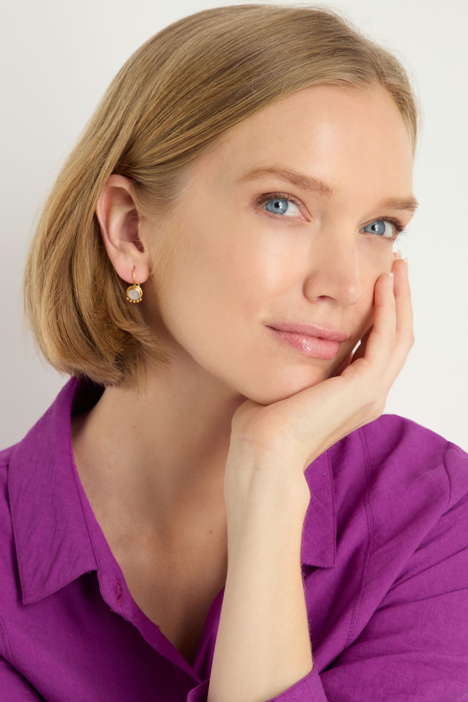 A woman with short blonde hair, wearing a purple shirt and Moonstone Earrings, rests her chin on her hand and looks slightly upward.
