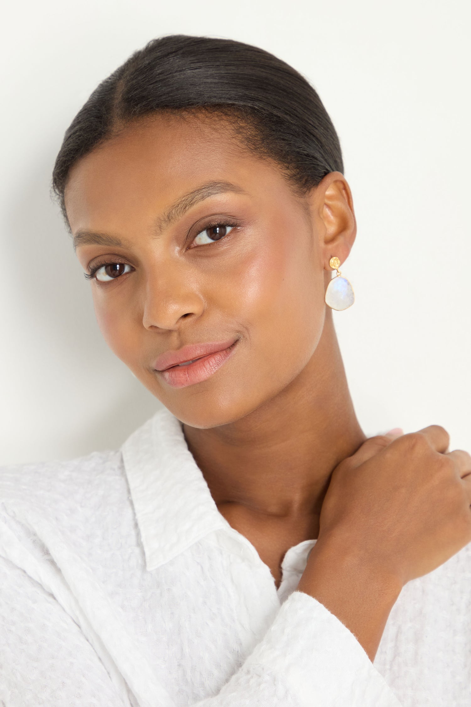 Woman with smooth dark hair pulled back, wearing a textured white top and elegant Hand Crafted Rainbow Moonstone Earrings, looking at the camera with a neutral expression.