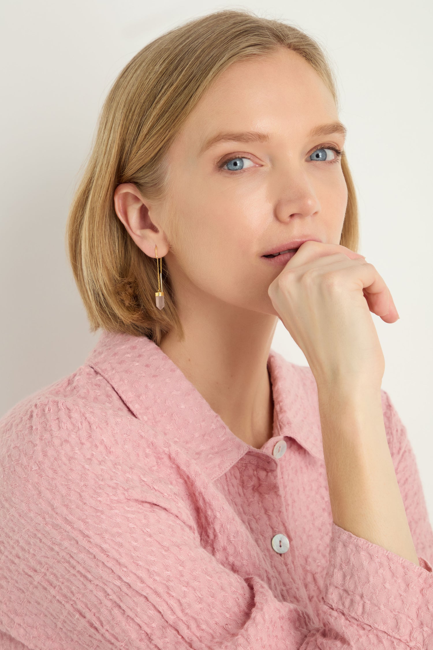 A woman with short blonde hair, wearing a textured pink shirt and Rose Quartz Crystal Drop Earrings, rests her chin on her hand while looking at the camera.