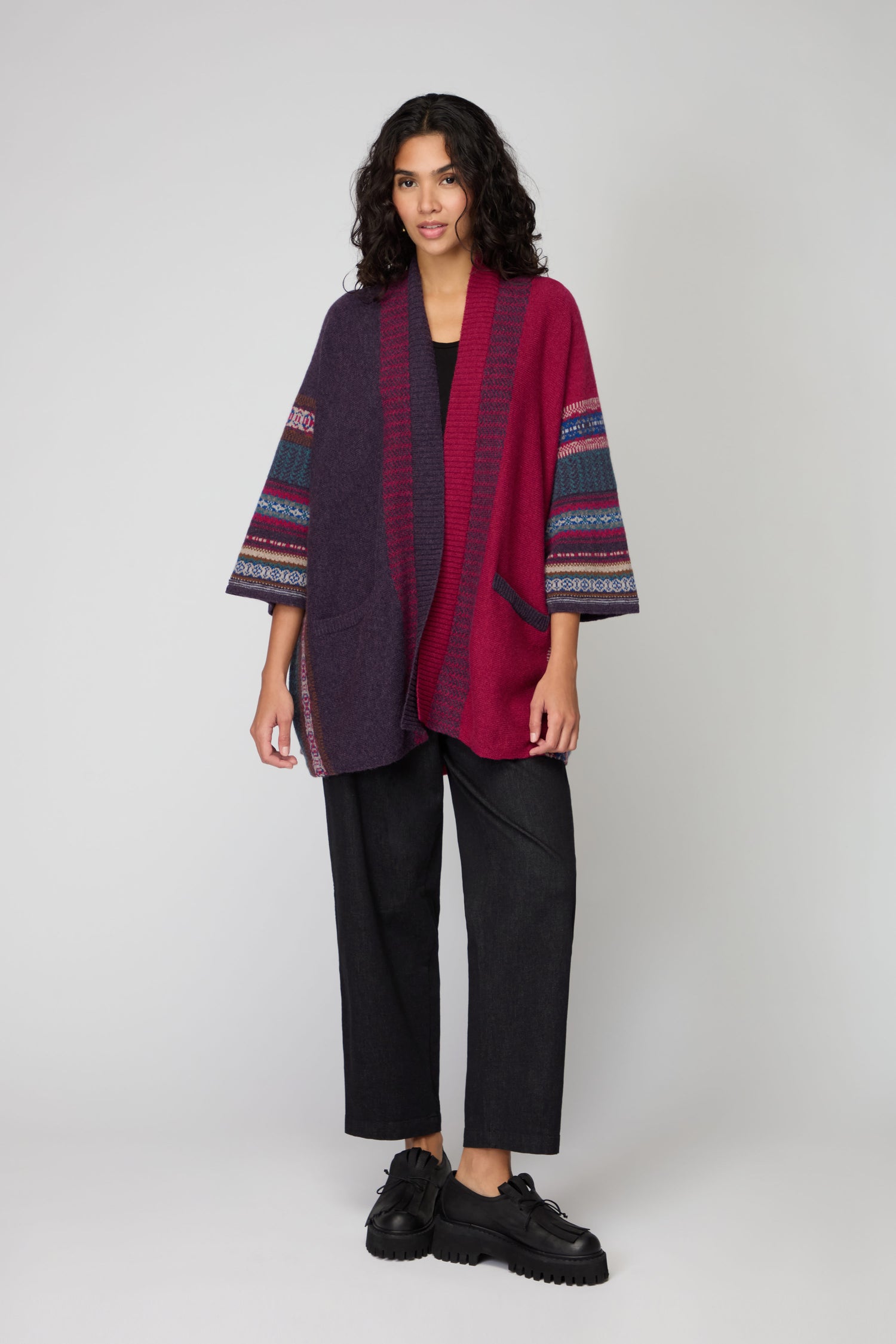 Woman standing against a plain background, wearing a Bramble Montrose Blanket Cardigan with patterned sleeves, a black top, wide-leg black pants, and black shoes.