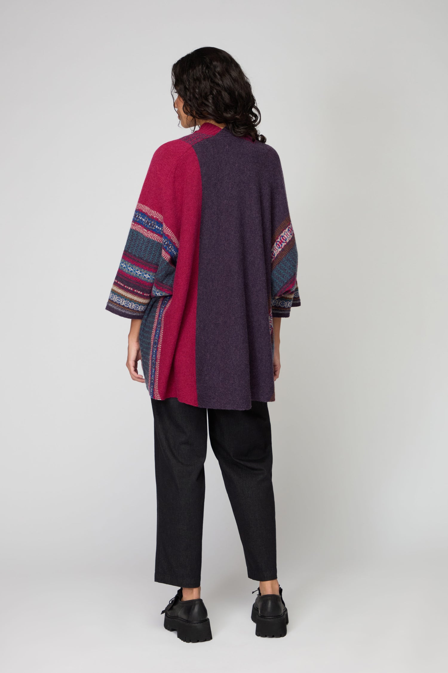 A person faces away, wearing the Bramble Montrose Blanket Cardigan with patterned sleeves, paired with black pants and shoes against a plain background.